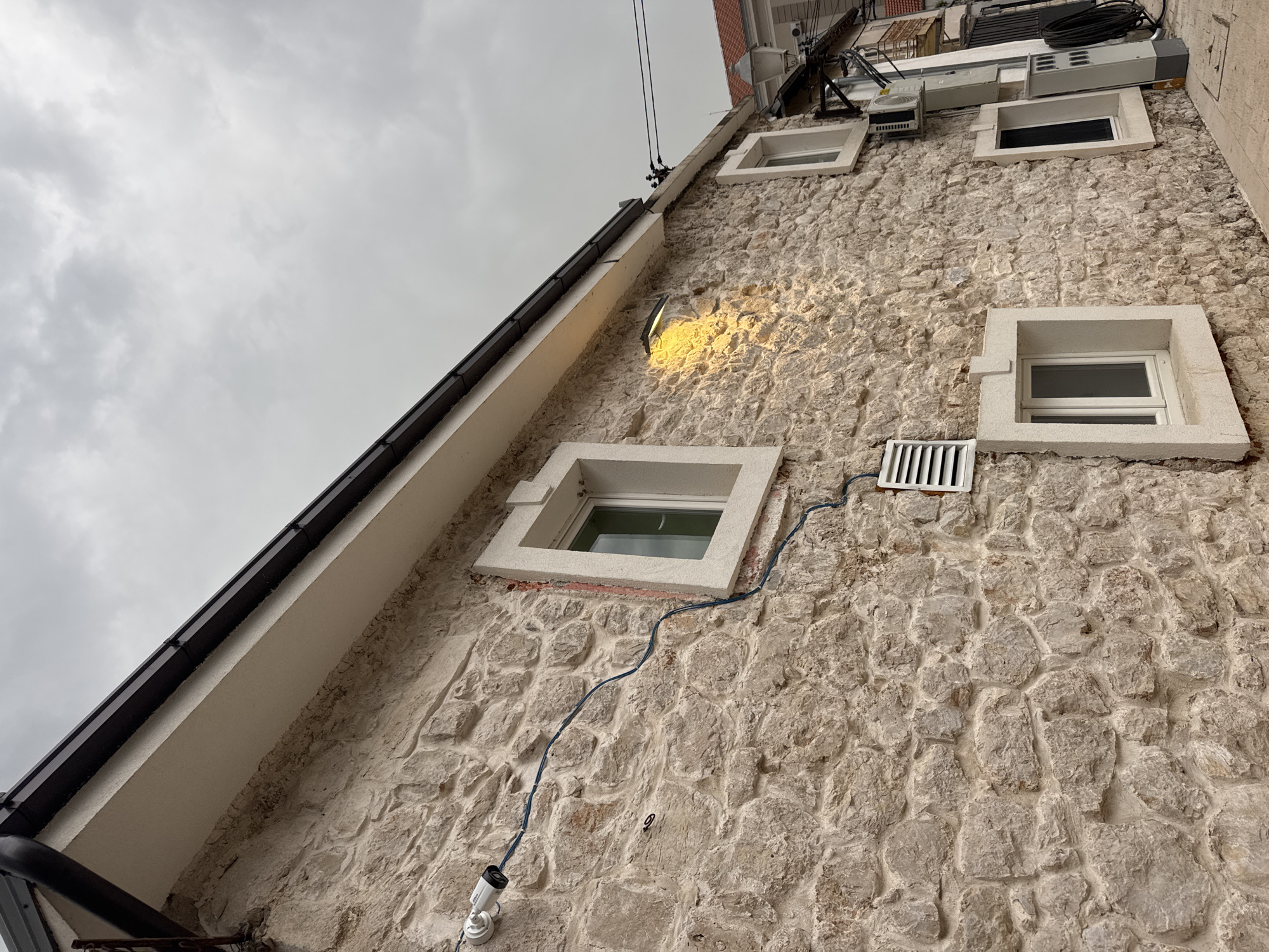 An exterior view of a stone building's facade in Trebinje, Bosnia and Herzegovina, under an overcast sky. The building features multiple windows with white frames, some of which appear to be closed. A security camera is mounted on the stone wall, with a blue wire running along the facade. A light fixture above and to the right of center illuminates a section of the stone wall with a warm yellow glow. Below the light, there are ventilation grates. Further down the wall, an outdoor air conditioning unit and electrical conduit are visible. The ground is paved, and a street lamp is present in the background. The stone masonry is irregular and weathered.