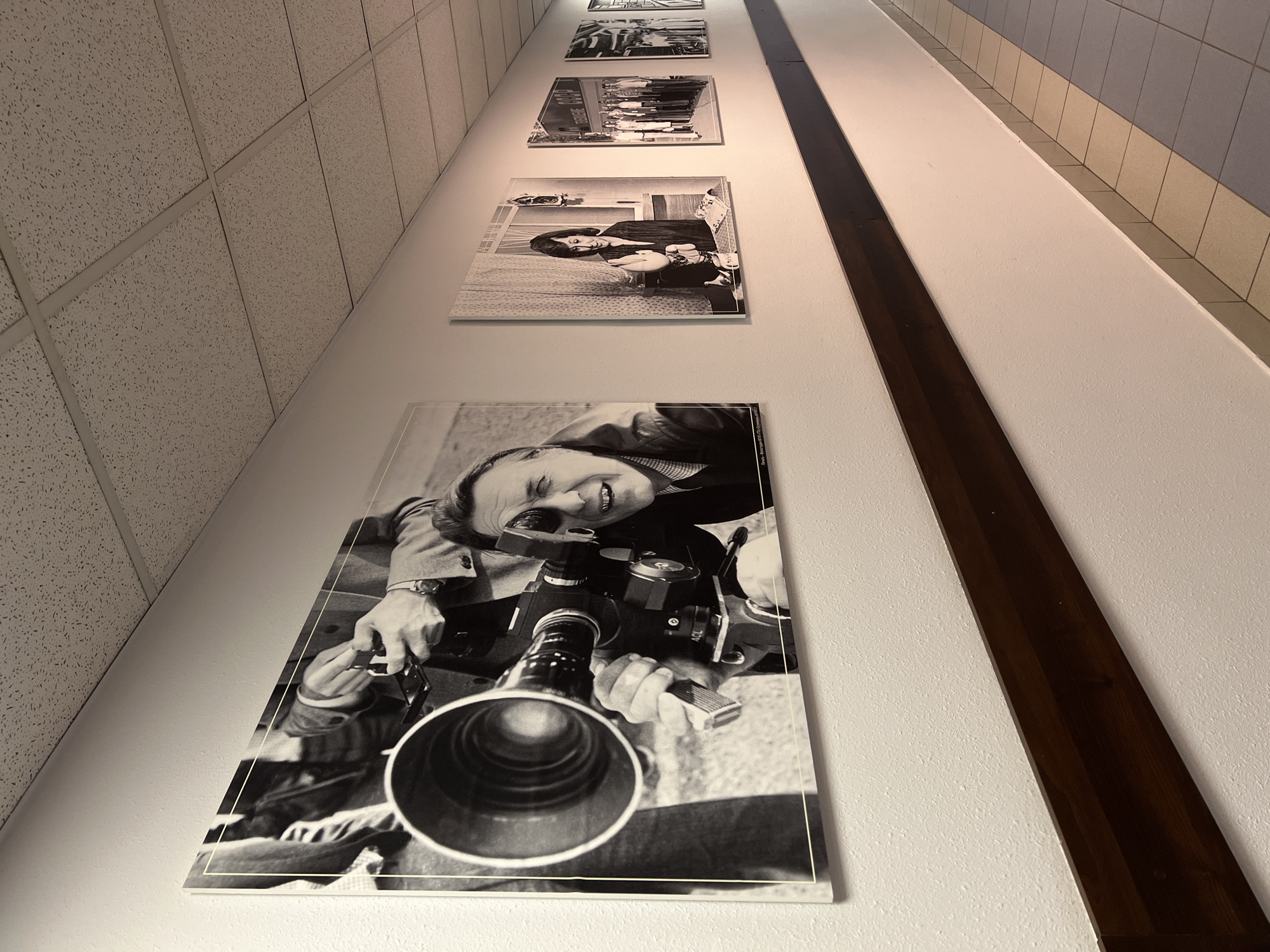 The image shows a series of black and white photographs displayed on a wall in an indoor space. The photos are hung in a vertical line, with the largest and most prominent photo featuring a man holding a camera with a large lens. The man in the photo is smiling and appears to be taking a picture. The other photos in the series appear to feature various people and scenes.
The wall is white, and a dark brown wooden strip runs vertically alongside the photos. The ceiling is visible in the upper left corner and appears to be a standard drop ceiling with rectangular tiles. The right side of the image shows a wall with light-colored square tiles above a darker beige section. The perspective is angled, giving a sense of depth to the hallway or corridor.
There is no information in the image to suggest that the location is in the city of Khimki, Russia.