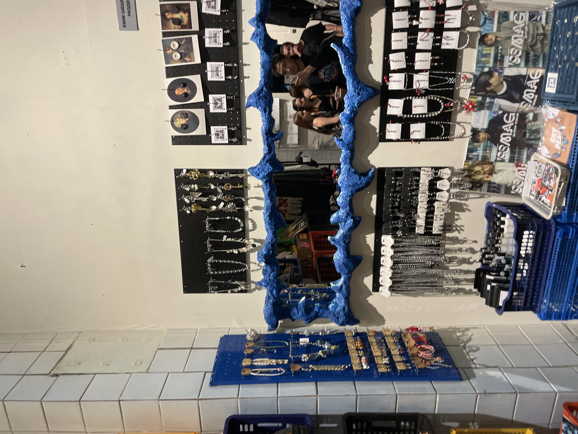 The image depicts an interior view of a retail space, likely a shop specializing in accessories. The scene is filled with various jewelry items displayed on pegboards and in baskets. On the left, a blue pegboard is adorned with necklaces, bracelets, and earrings. To the right, several black pegboards showcase an assortment of necklaces. In the center, a mirror with a vibrant blue, textured frame reflects a group of young adults, one of whom is taking a selfie. The background reveals stacks of blue plastic crates, some containing packaged items and bottles, suggesting inventory or display storage. Posters featuring young men in fashionable attire are visible at the bottom, further indicating a retail environment focused on youth culture or fashion. The walls are partly covered with white square tiles. The overall impression is that of a trendy boutique or market stall. The specific location in Almaty, Kazakhstan, cannot be determined from the visual information provided.