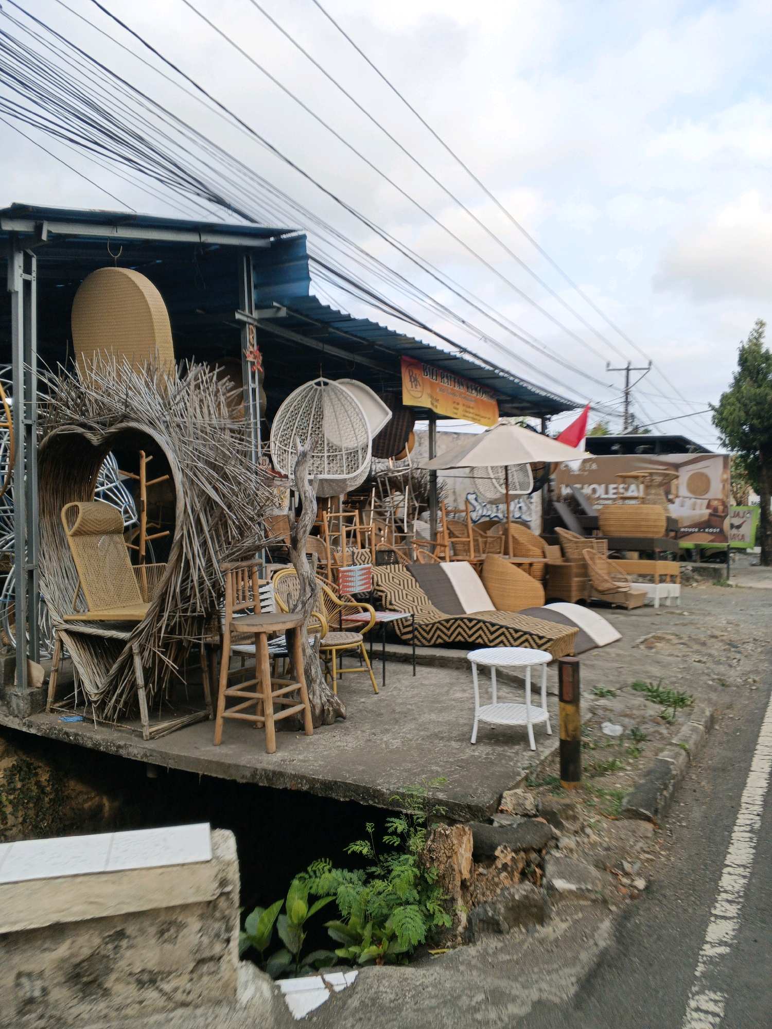 This is a medium-shot, eye-level, outdoor, daytime, slightly overcast photograph of a furniture store in Sukawati, Indonesia. The store is filled with rattan furniture, including chairs, loungers, and decorative items. In the foreground, a large, heart-shaped rattan sculpture is prominently displayed. A yellow rattan chair is placed inside the sculpture. To the right of the sculpture, there is a wooden stool and a white rattan hanging chair. Further back, more rattan furniture is visible, including loungers with patterned cushions, beanbag-like chairs, and tables. An umbrella stands partially open, providing shade. The store has a blue awning and is situated along a paved road. Electrical wires are strung across the top of the image. The ground in front of the store is concrete, and there appears to be a small channel of water or ditch on the left side. The overall scene depicts an outdoor retail environment selling natural-fiber furniture in a tropical setting.