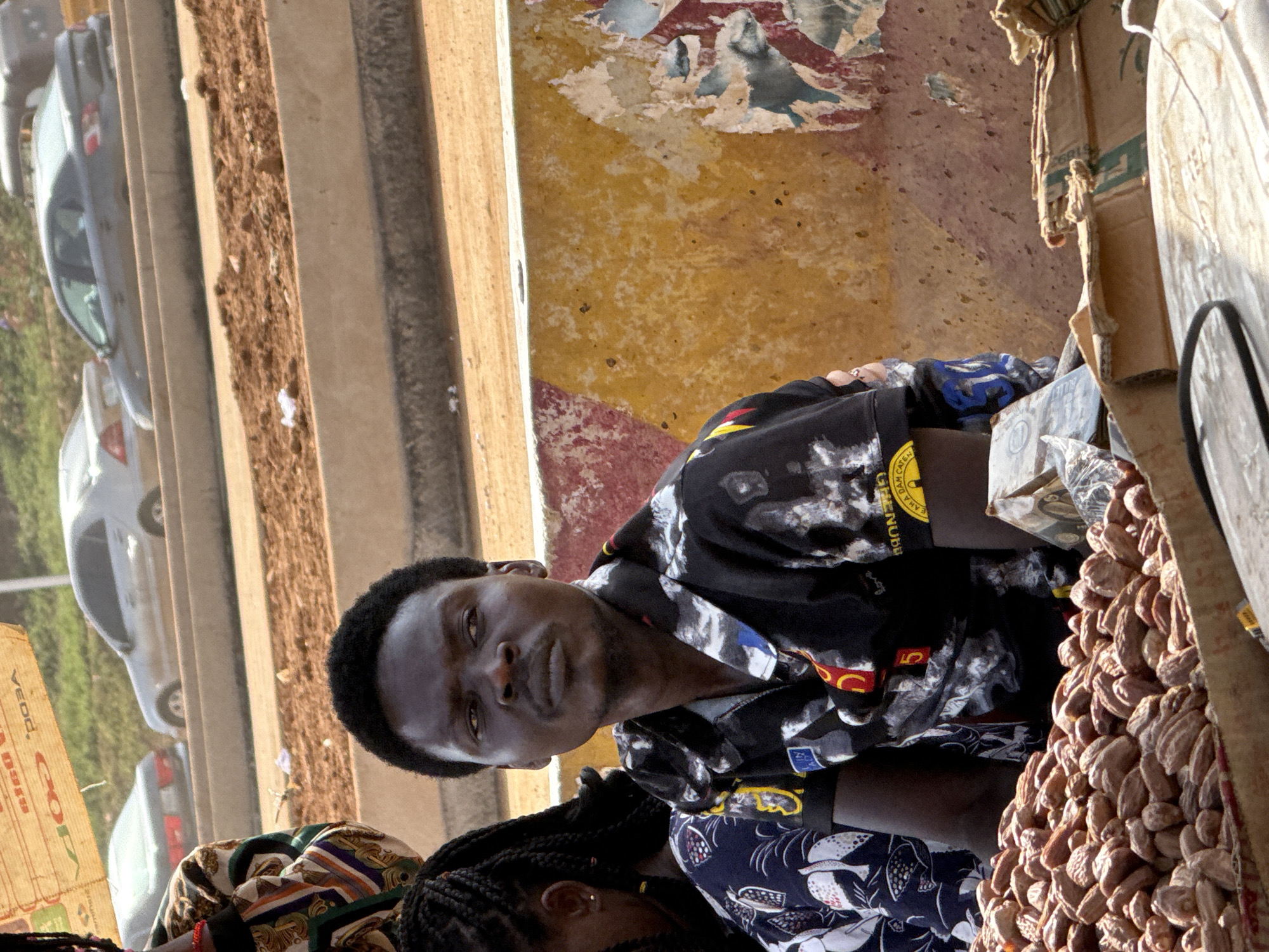 A medium, slightly-high-angle, indoor shot shows a man and a woman seated in front of a pile of nuts. The man is African, dark-skinned, with close-cut black hair. He is wearing a black shirt with a white, blue, and red pattern. A wristband of yellow with black text is visible on his right arm. He looks directly at the camera. A woman sits next to him, slightly lower in the frame. Only her head and shoulders are visible. She has dark skin and black, braided hair. She wears a blue and white top. The background shows a concrete structure. Cars are visible outside. Part of a cardboard box is visible at the right edge of the frame. Part of a sign is visible at the left edge of the frame. The location is suspected to be in Abuja, Nigeria.