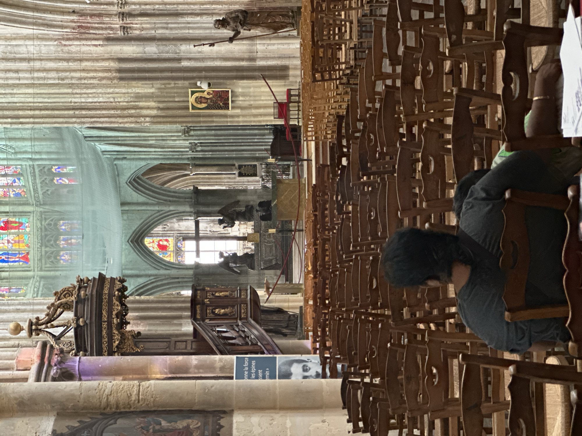 The image depicts the interior of the Saint-Jacques church in Dieppe, France. In the foreground, a person is seated with their back to the camera, facing rows of empty wooden chairs. The church's architecture is visible, including tall columns, arched windows with stained glass, and a pulpit. Further back, a large crucifix is present, flanked by statues of angels. The overall scene conveys a sense of a quiet and solemn religious space.