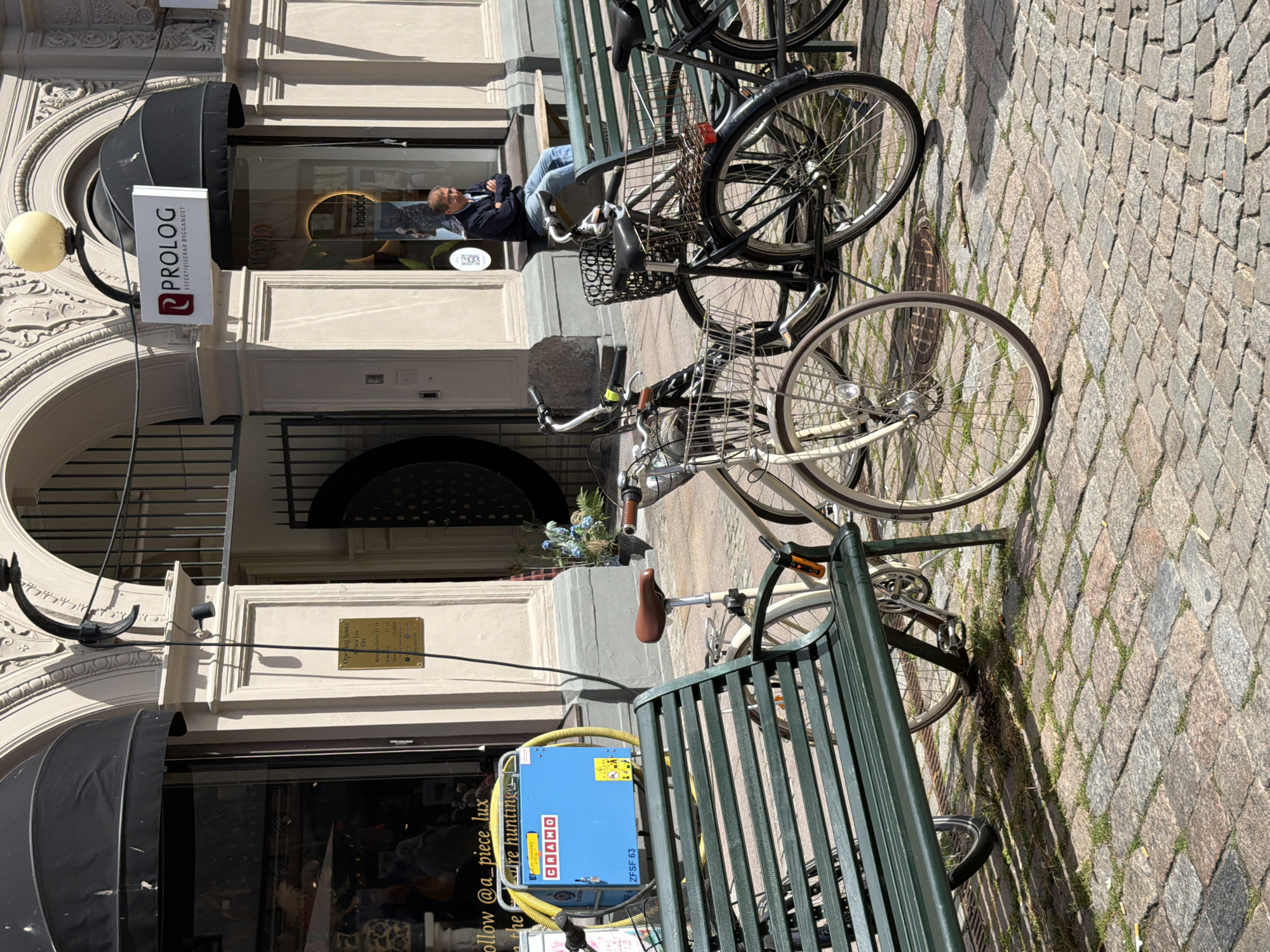 The image shows a street scene in Malmö, Sweden, in front of a building with awnings and arched entrances. In the foreground, a green park bench is placed on a cobblestone street. Several bicycles are parked next to the bench. One bicycle, partially visible on the left, is cream-colored with a basket. A black bicycle with a basket and a red reflector is parked in the center. Further to the right, another black bicycle is visible.
Behind the bench and bicycles, a blue electrical device with yellow hoses is placed on the ground. The device has "CRAMO" written on it. Above the device, a sign indicates "follow @a_piece_lux" and "are hunting".
A man is seated on a bench further back in the scene, in front of a shop with a sign that reads "PROLOG EFFEKTIVISERAR BYGGANDET". The man is wearing a dark jacket and blue jeans and has his arms crossed. Another shop window displays "headon". The architecture of the building features ornate detailing and arched doorways. The scene is bathed in sunlight, creating shadows on the cobblestones.