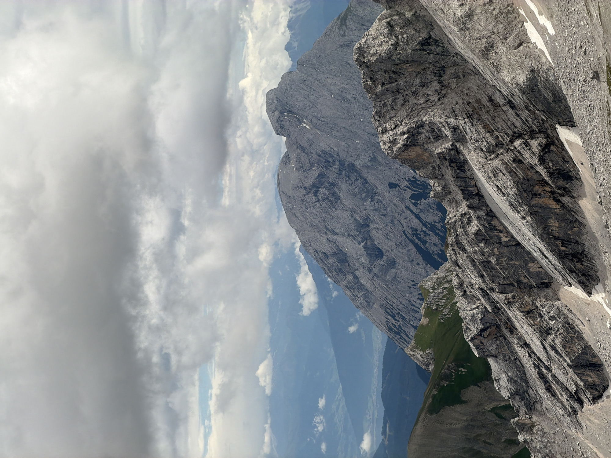 This is an image of a mountainous landscape in Wildermieming, Austria, under a cloudy sky. The foreground features a rocky, grey mountain slope with patches of snow and scree. Further back, a large, rugged grey mountain dominates the center of the frame, its peak partially obscured by clouds. In the distance, layers of hazy blue mountains recede into the background, with hints of a valley and some vegetation visible in the far distance. The clouds are thick and grey, with some lighter patches indicating breaks in the cloud cover. There are no people or man-made objects visible in the image.