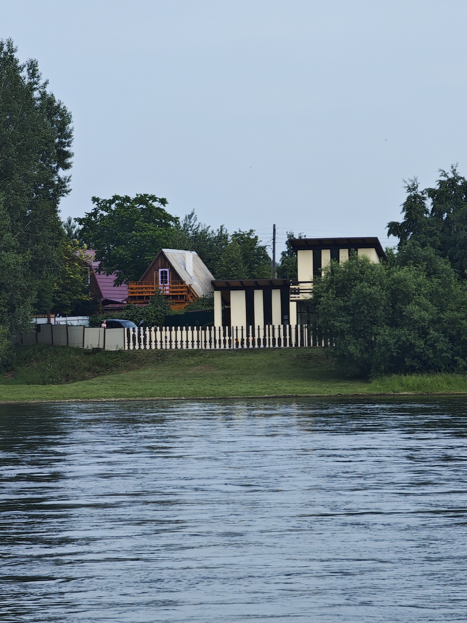 The image depicts a waterfront scene in the city of Sosnovoborsk, Russia. The foreground is dominated by a wide body of water, likely a river, with visible ripples and reflections. The middle ground features a grassy bank separating the water from a residential area. Several houses are visible, characterized by their varied architectural styles. To the left, a house with a red roof and light-colored siding is partially obscured by trees. Adjacent to it is a house with a steep, light-colored roof and a wooden balcony, featuring a small purple window. A white picket fence runs along the property line. A dark-colored car is parked near this house, with its driver's side door open. Further to the right, another modern-looking structure with alternating dark and light vertical panels is situated. This building also has a balcony and is surrounded by lush green trees. A light-colored fence encloses the properties. The background consists of a pale, overcast sky. The overall impression is a tranquil, suburban riverside setting.