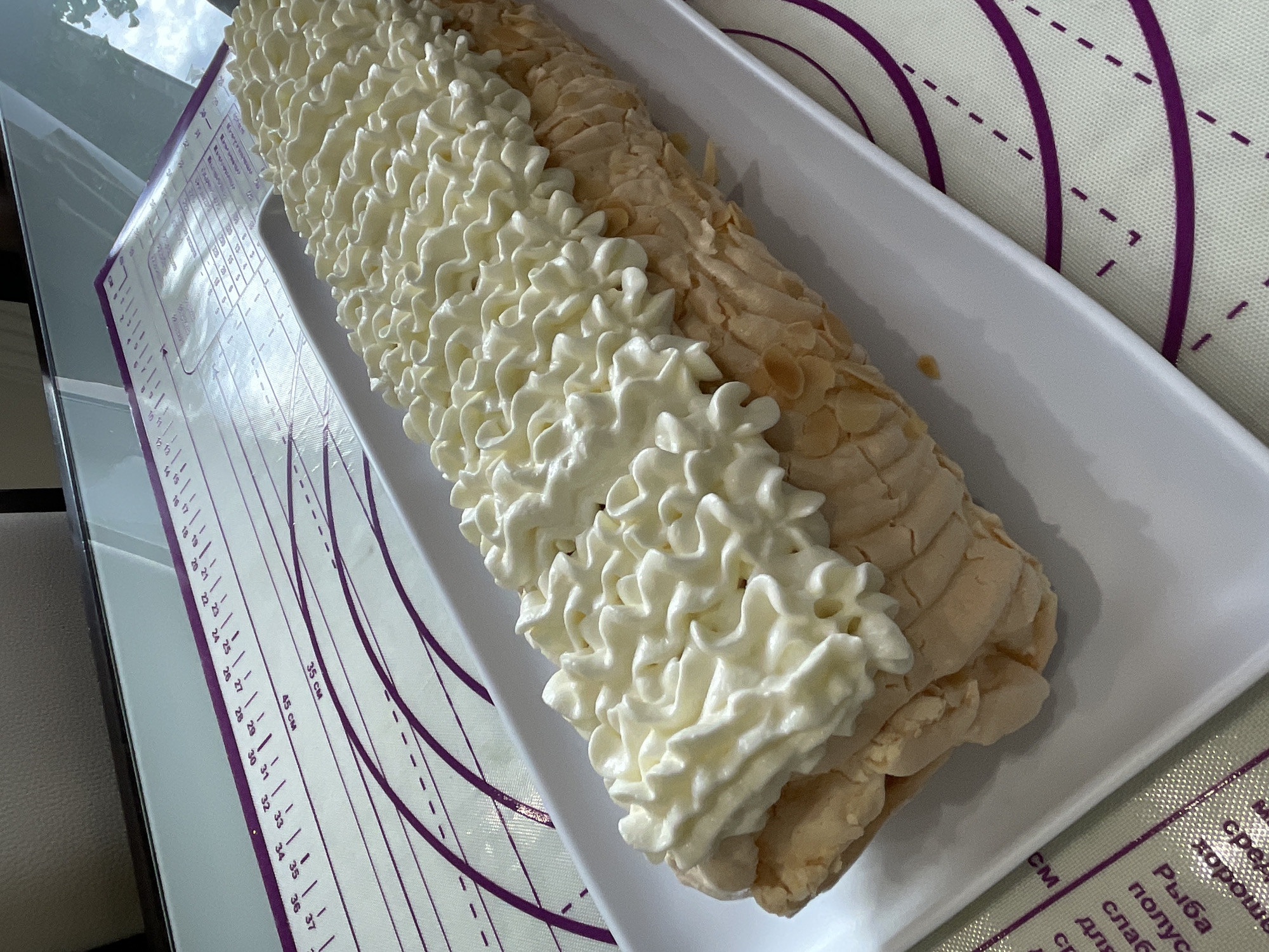 The image shows a white rectangular platter holding a rolled dessert. The dessert appears to be a roulade with a light brown, textured exterior, possibly meringue or cake, covered with white whipped cream piped in a decorative pattern along the top. The platter is placed on a purple and white baking mat with measurement markings. The background is out of focus but suggests an indoor setting with a reflective surface, possibly a table. There are no people or animals visible in the image. The scene is static and appears to be a close-up product shot of the dessert. The location is not identifiable within the city of Saint Petersburg, Russia.