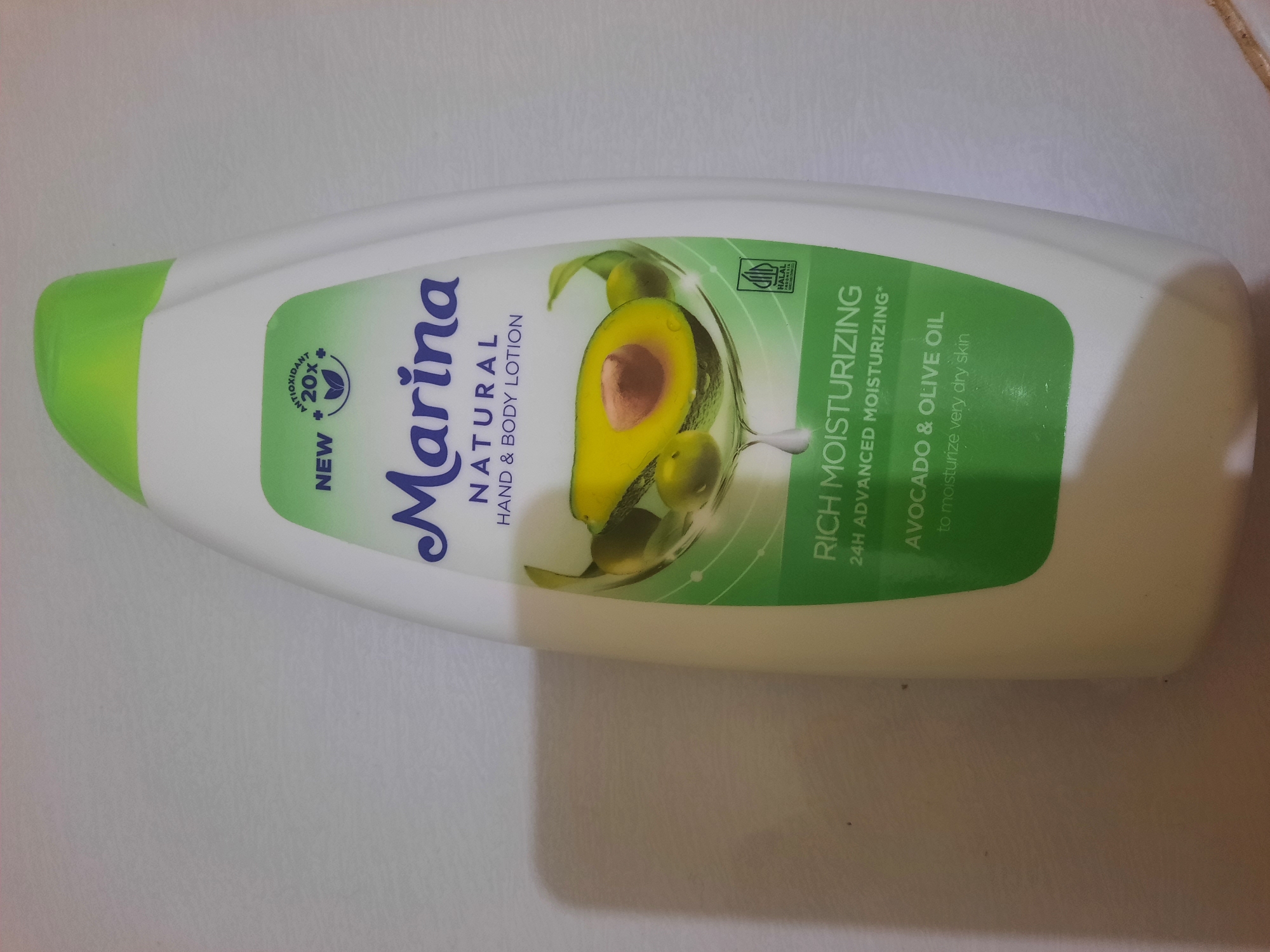 A white plastic bottle of Marina Natural Hand & Body Lotion is shown against a white background. The bottle has a green cap and a green label with avocado and olive illustrations. Text on the label includes "NEW," "ANTIOXIDANT 20X," "Marina," "NATURAL," "HAND & BODY LOTION," "RICH MOISTURIZING," "24H ADVANCED MOISTURIZING," "AVOCADO & OLIVE OIL," and "to moisturize very dry skin." A small, square HALAL logo is visible on the lower right of the label. The bottle is standing upright, and a shadow is cast to the left. There is no indication of Lhokseumawe, Indonesia, in the image or its context.