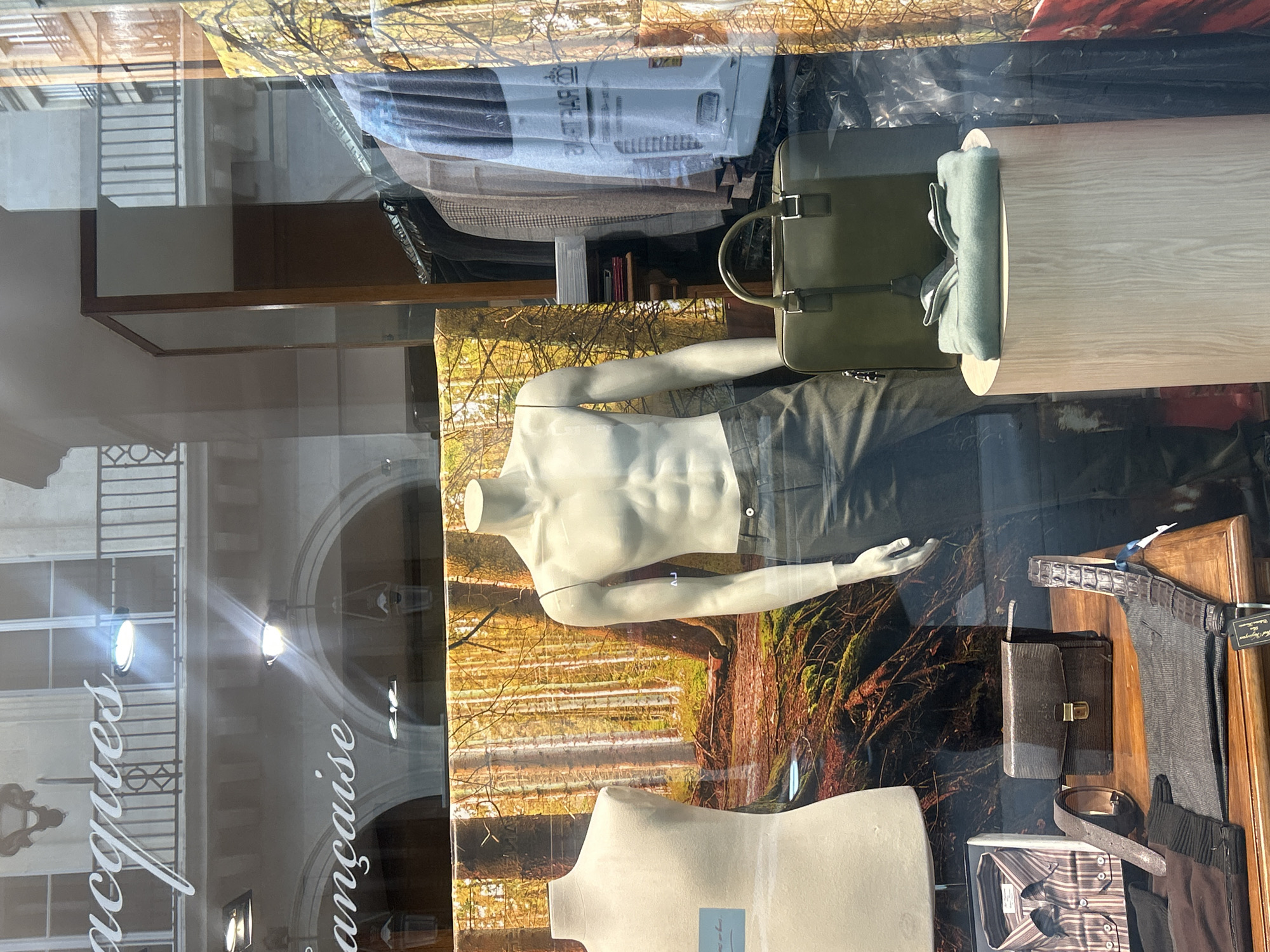 A retail display is seen in a window of a business displaying men's clothing and accessories. Two white, torso-only mannequins are positioned against a backdrop of an autumnal forest scene. The upper mannequin wears dark trousers. A green bag sits atop a light-colored wooden display stand to the side. Additionally, an assortment of accessories, including belts, a tie, and what appears to be a leather portfolio are visible alongside the trousers. The window reflection reveals building facades and architecture from across the street. The business name, "Jacques Française" is visible on the window.