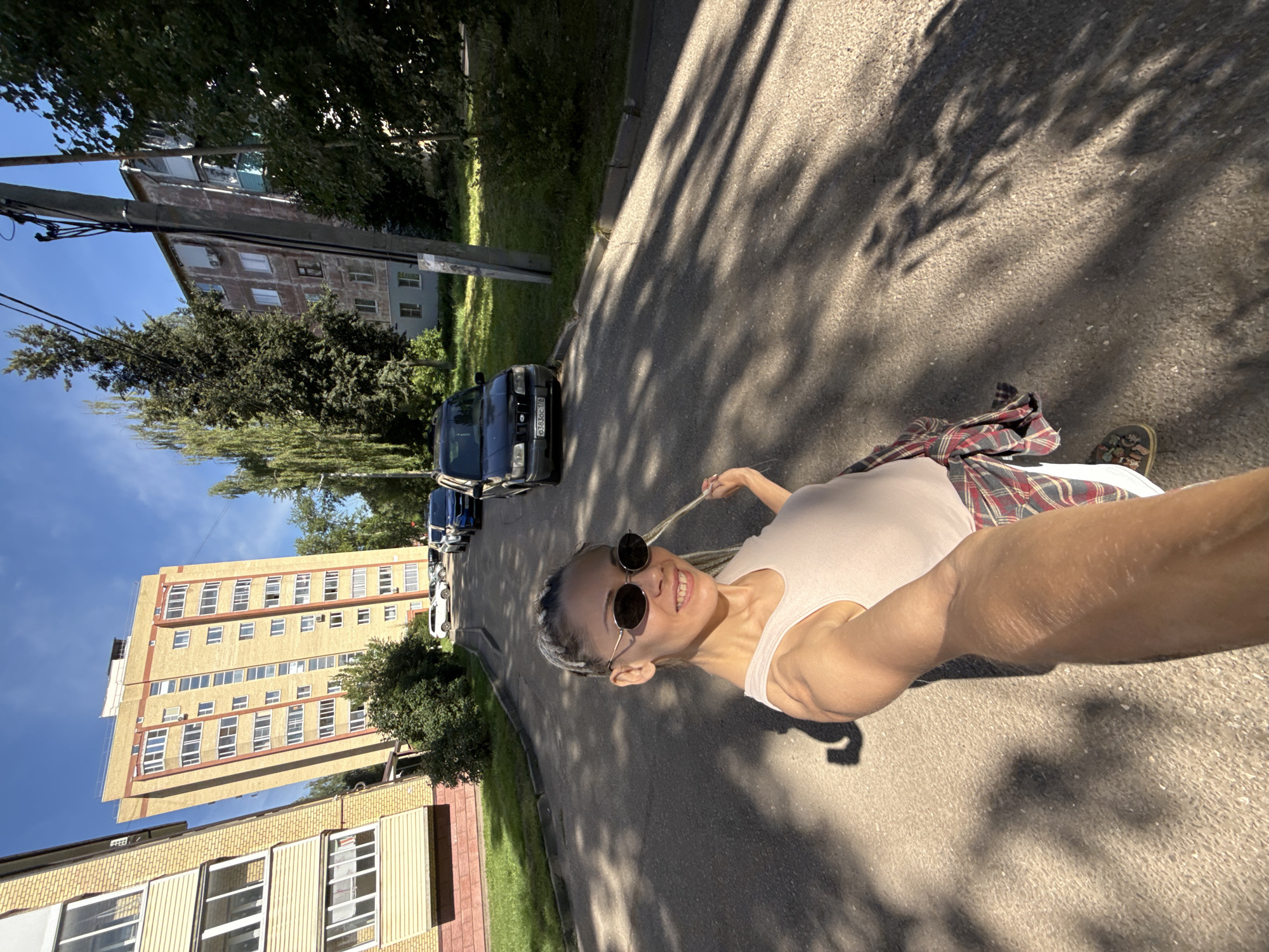 A young woman with braided hair and sunglasses takes a selfie outdoors on a sunny day. She is wearing a light-colored tank top and a plaid shirt tied around her waist. The background shows a street with several parked cars, apartment buildings, and trees. The architecture of the buildings suggests an urban setting. The scene is likely in Rusanovo, Russia, as indicated by the style of the buildings and the presence of cars. The shadows on the ground indicate a bright, sunny day.