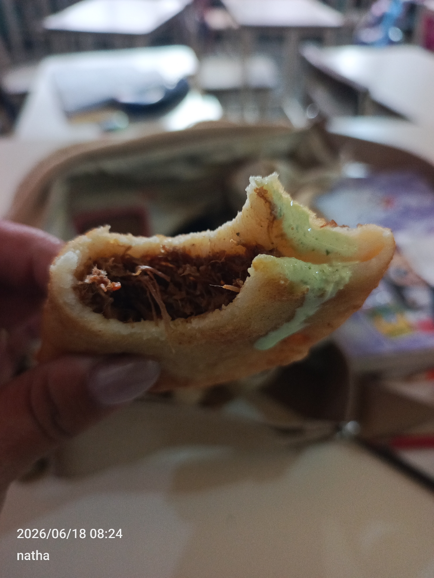 The image shows a close-up of a person's hand holding a piece of food, possibly an arepa, which has been bitten into. The food appears to be fried or baked dough, filled with shredded meat, and topped with a light green sauce, likely avocado or a similar creamy condiment. The background is blurred, suggesting an indoor setting, perhaps a classroom or a casual dining area, with tables and chairs visible. The lighting indicates it might be daytime. Text overlay on the bottom left corner reads "2026/06/18 08:24" and "natha". The overall impression is that of a quick snack being enjoyed, possibly on the go, given the casual way it's being held and the focus on the food itself. The location context provided is San Sebastián, Venezuela.