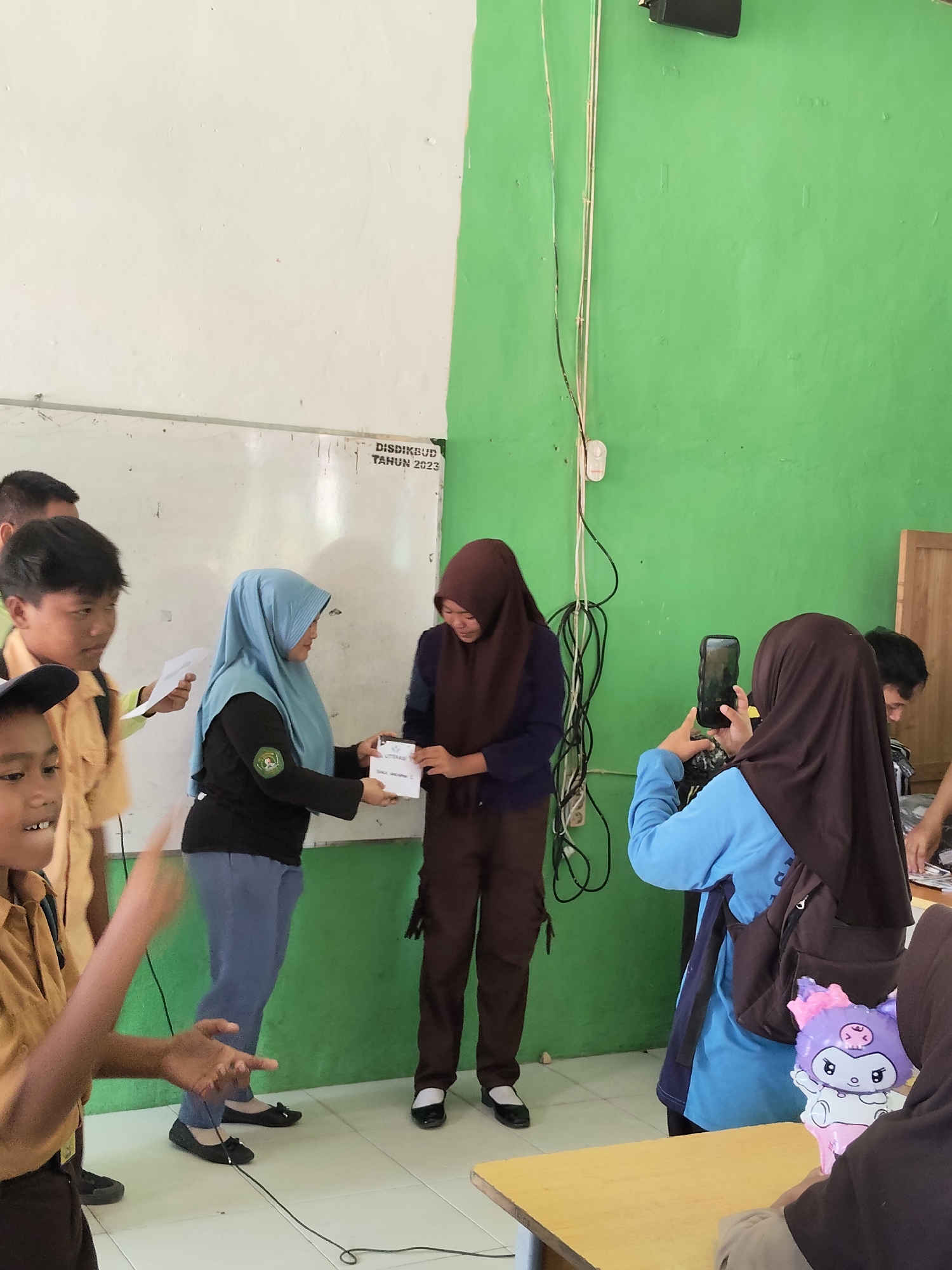 In a room with white and green walls, a group of people, likely students, are gathered. Two young women in hijabs are at the center of the activity. One, wearing a blue hijab, is handing a small item, possibly a certificate or award, to another woman in a brown hijab. The second woman is wearing a dark blue shirt and brown pants. She is looking down at the item being exchanged. A younger boy in a tan uniform with a black cap is in the foreground on the left, looking towards the center with his mouth open and hands in a gesture that suggests excitement or talking. Another boy, also in a tan uniform, is standing behind him, looking towards the center and holding a piece of paper. To the right, a young woman in a brown hijab is holding up a smartphone, seemingly recording the event. She is wearing a blue long-sleeved shirt and has a backpack with a cartoon character charm. The room appears to be a classroom or community space, with a whiteboard visible on the left wall containing text that reads "DISDIKBUD TAHUN 2023". Electrical cords are hanging down the green wall. A wooden table is present in the foreground on the right. The scene suggests an award ceremony, presentation, or school event. The location is identified as Lori, Indonesia.