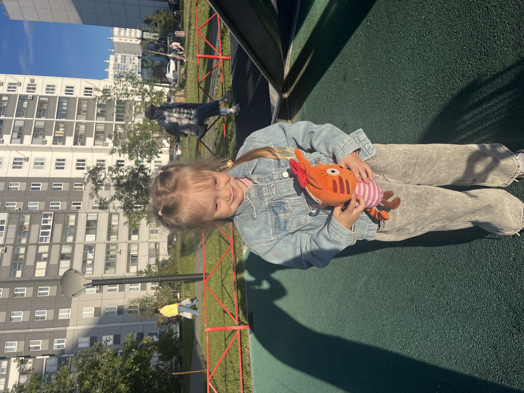 A young girl, approximately 4-6 years old, stands centered in the foreground on a textured green rubber playground surface. She has light blonde hair styled in two pigtails, each secured with a light-colored hair elastic that resembles a flower. She wears a light blue denim jacket over a pink top and light grey wide-leg pants. Her feet are partially visible, showing white sneakers with red accents. She holds an orange plush toy with white and pink stripes on its belly, black stripes on its body, large white eyes with black pupils, and two orange ears. The girl is smiling slightly, looking downwards and to her left.
Strong directional sunlight from the right casts a prominent, long, dark shadow of a person (likely the photographer) directly in front of the girl, extending from the bottom center of the frame towards the upper left. Another dark shadow, possibly from a playground structure, is visible on the green surface to the girl's right.
In the midground, a red metal fence runs horizontally, separating the playground from a grassy area and a paved path. Beyond the fence, several individuals are visible. To the left, a person in a bright yellow long-sleeved shirt and blue pants walks away, carrying two white bags. To the right, another individual in a plaid shirt and blue jeans sits on a low structure, possibly a bench or part of the playground. Further back, two more individuals are seated near a light-colored trash receptacle. Several trees and green bushes are scattered throughout the midground. A tall, grey street lamp with a flat, circular top stands to the left of the center.
In the background, multiple large residential apartment buildings dominate the skyline. These buildings are predominantly light grey and white, featuring numerous rectangular windows, some of which have small air conditioning units mounted beneath them. The sky above is clear and blue. Several cars are parked intermittently behind the buildings and trees. The scene depicts an urban residential environment during daylight hours.