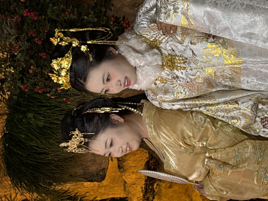 The image depicts two adult East Asian women, dressed in elaborate traditional Chinese garments, posing in an outdoor, landscaped setting during nighttime in Yuzhong District, China. The specific location within Yuzhong District is not visually identifiable but includes dense green foliage and rock formations illuminated by warm artificial light.
The woman on the left, positioned slightly behind the woman on the right, wears a lustrous gold-colored robe with long sleeves. Her dark hair is styled upwards, adorned with a golden crown-like headpiece that includes intricate designs and dangling elements. She holds a white, fan-like object with a scalloped edge and pearl-like embellishments in her left hand, visible at waist level. Her expression is a smile, and she is looking towards the right side of the frame.
The woman on the right wears a white robe extensively embroidered with gold patterns, depicting phoenix and floral motifs. A white, fluffy faux fur collar surrounds her neck, below which a prominent gold pendant with amber-colored dangles is visible. Her dark hair is also styled upwards, featuring a golden headpiece with floral elements and cascading chains. A pearl earring is visible on her right ear. She is smiling and looking towards the right.
The background consists of dark green vegetation, including what appear to be bushes and vines, with clusters of small red flowers or berries visible on the right. Yellowish-brown rockwork is present on the left side of the background. The scene is brightly lit by artificial sources, creating a warm glow.