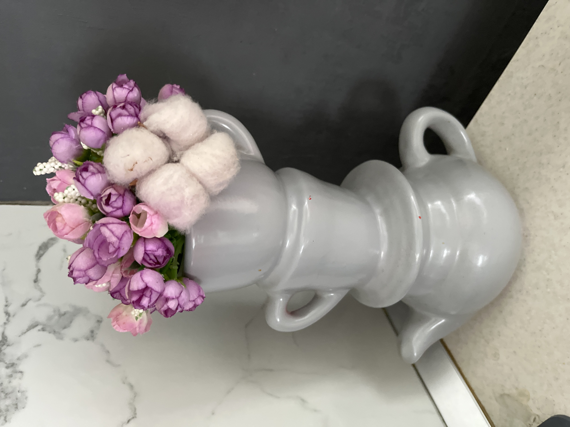 A still life composition features a stack of three gray ceramic tea-themed objects: a teapot at the base, topped by two teacups, one inverted. A bouquet of artificial pink and purple flowers with small white accents and fluffy white decorative elements is arranged within the topmost teacup. The objects are placed on a speckled, light-colored countertop. In the background, a section of a white marble-patterned wall is visible to the left, and a dark gray wall occupies the right side. The scene is brightly lit. The image does not contain any information about the city of Saratov, Russia, or any individuals.