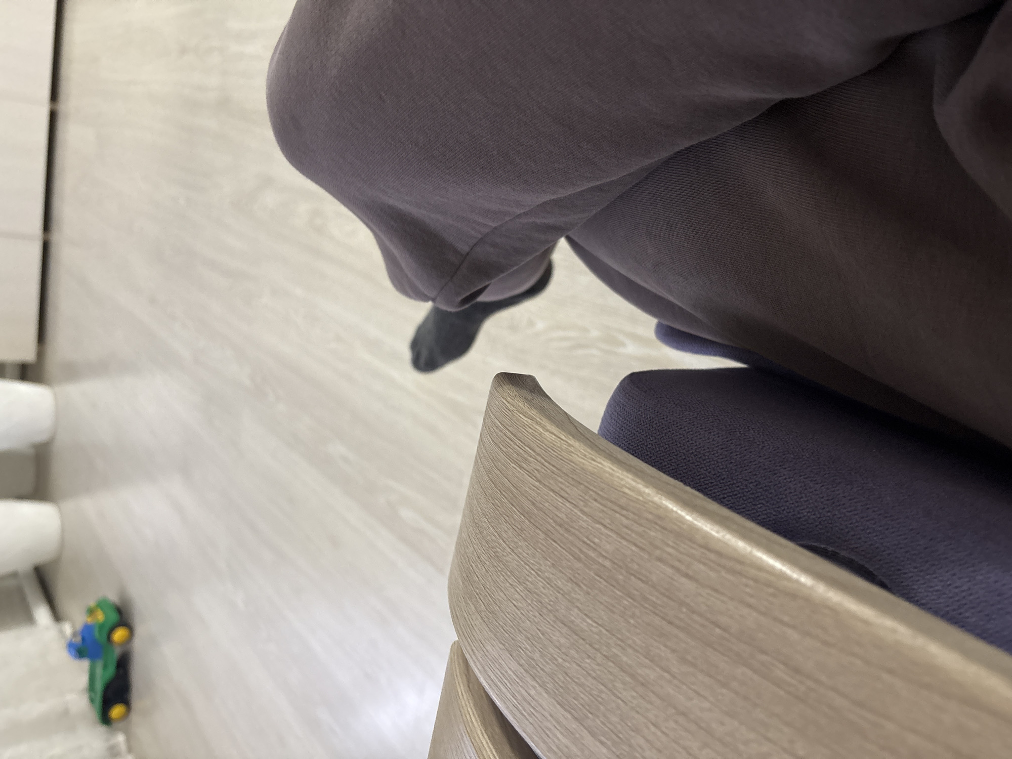 The image shows a person sitting on a chair, with a gray sock visible. The chair is wooden with a blue seat. In the background, a toy and a white structure, possibly a bathroom fixture, are visible on a light-colored floor. The location in the city of Ферма, Russia cannot be verified from the image.