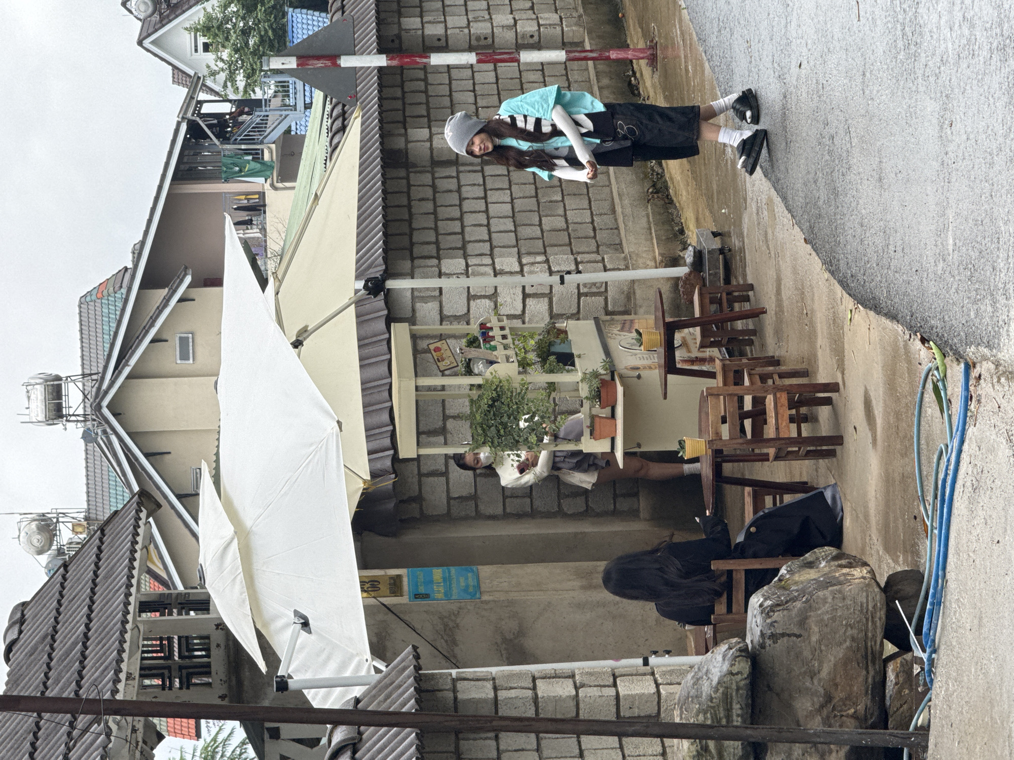 This image captures an outdoor cafe scene nestled along a paved pathway in a residential area, likely in Lâm Viên - Đà Lạt, Vietnam, given the architectural style of the surrounding houses. The sky appears overcast, suggesting a daytime shot with soft, diffused light.
Three individuals are present. On the right side of the path, a young woman stands, wearing a light blue short-sleeved top over a white long-sleeved shirt, a dark skirt, white socks, and black sandals. She completes her outfit with a grey beanie and appears to be holding a small bag or item in her hands, looking slightly towards the cafe area with a faint smile.
In the mid-ground, an open white outdoor umbrella provides shade for a seating area with multiple wooden tables and chairs. Behind one of these tables, a person is seated, partially obscured by a plant-laden shelf, wearing a white top and a face mask. Further to the left and slightly to the foreground, another person is seated with their back to the camera, showcasing long dark hair and wearing a dark jacket or top.
Notable objects include a red and white striped barrier post near the standing woman, large grey rocks positioned on the ground, and a blue hose coiled nearby. The background features several multi-story houses with brown and colorful tiled roofs, some with visible balconies and structures like water tanks or antennas on their rooftops. A blue sign with white text, partially readable as "TRAT LAM HOP" or similar, is affixed to a wall behind the cafe's plant shelf. The ground appears slightly damp in places, hinting at recent rain or general moisture. The overall ambiance is relaxed and casual.