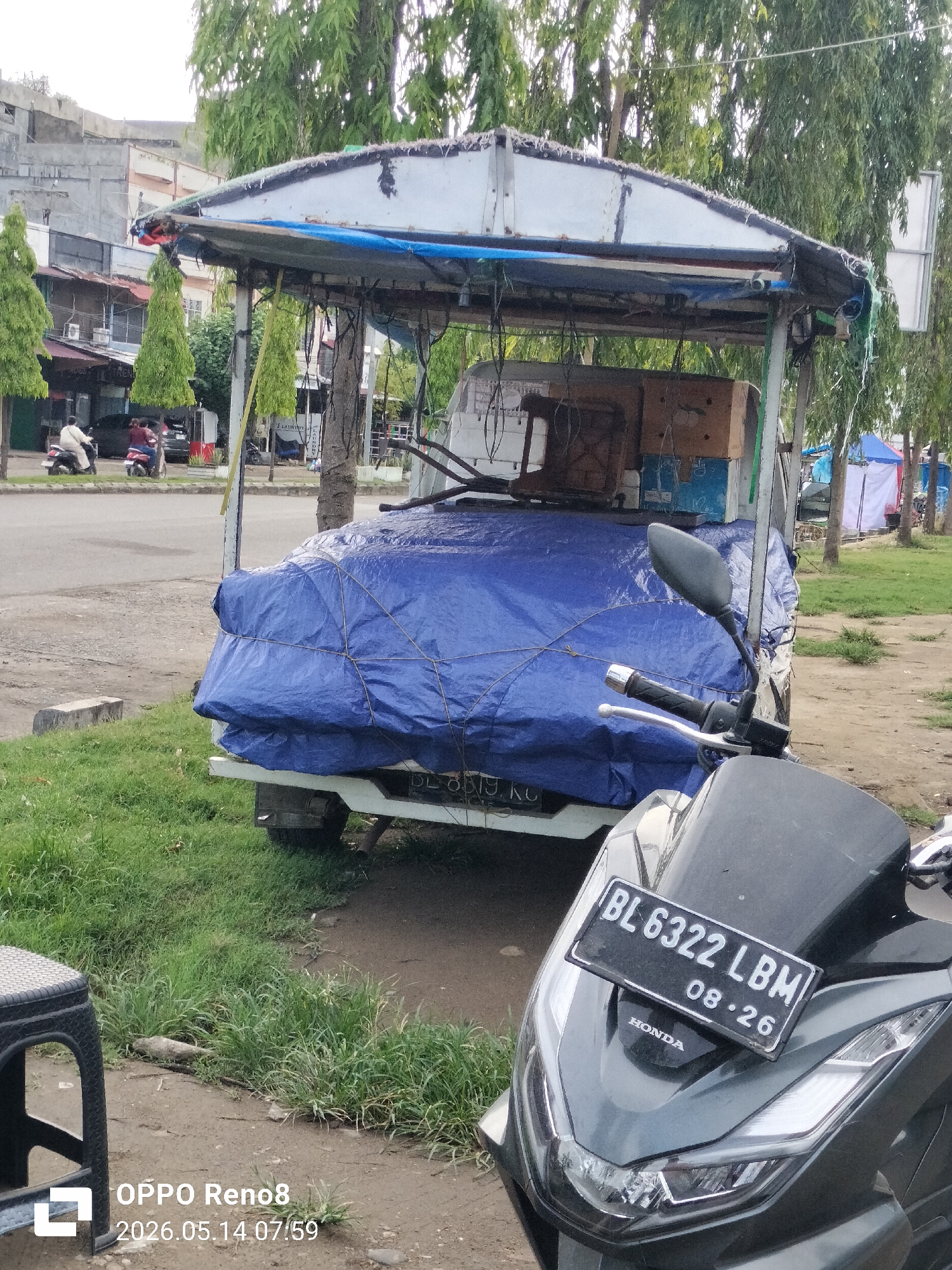 The scene depicts a street in Lhokseumawe, Indonesia. In the foreground, a parked Honda motorcycle with the license plate "BL 6322 LBM" is visible. Behind the motorcycle, a white truck is covered with a large blue tarp and has a makeshift canopy structure. The back of the truck reads "819 KG" in black lettering. On the left side of the image, two individuals on motorcycles are visible on a street. Buildings are located in the background behind trees. In the lower left corner, a black plastic stool is seen. The scene appears to be outdoors in daylight. The timestamp "2026.05.14 07:59" is watermarked in the bottom left corner.