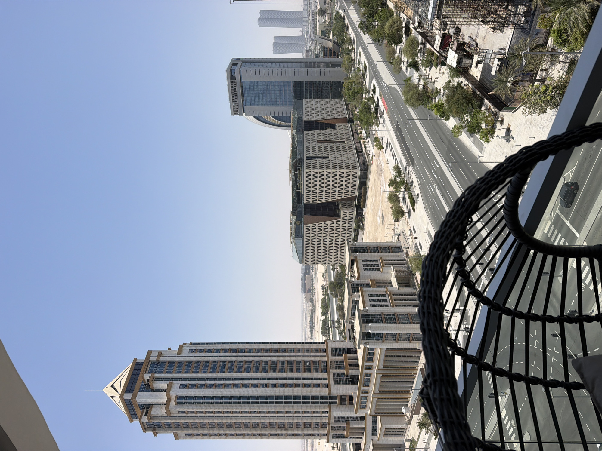 The image shows a cityscape dominated by modern architecture under a clear blue sky. In the foreground, a portion of a dark woven chair is visible, suggesting a viewpoint from a balcony or high-rise. The cityscape features several distinct buildings. To the left, a tall, multi-story building with a pyramidal top is prominent, characterized by its beige facade and numerous vertical windows. Adjacent to it is a lower, multi-winged structure with similar beige tones and extensive glass facades. Further into the background, a cluster of buildings includes a large structure with a distinctive curved roofline and another tall, rectangular building with a dark facade and a prominent glass-fronted upper section. In the distance, two identical, tall, slender buildings stand out against the horizon, suggesting a more densely built urban area. A wide, multi-lane road with clear lane markings runs through the scene, flanked by sidewalks and sparsely planted trees. The ground in many areas appears to be undeveloped, with sandy soil and some construction sites visible, indicated by scaffolding and building materials. The overall impression is one of a modern, developing urban environment.