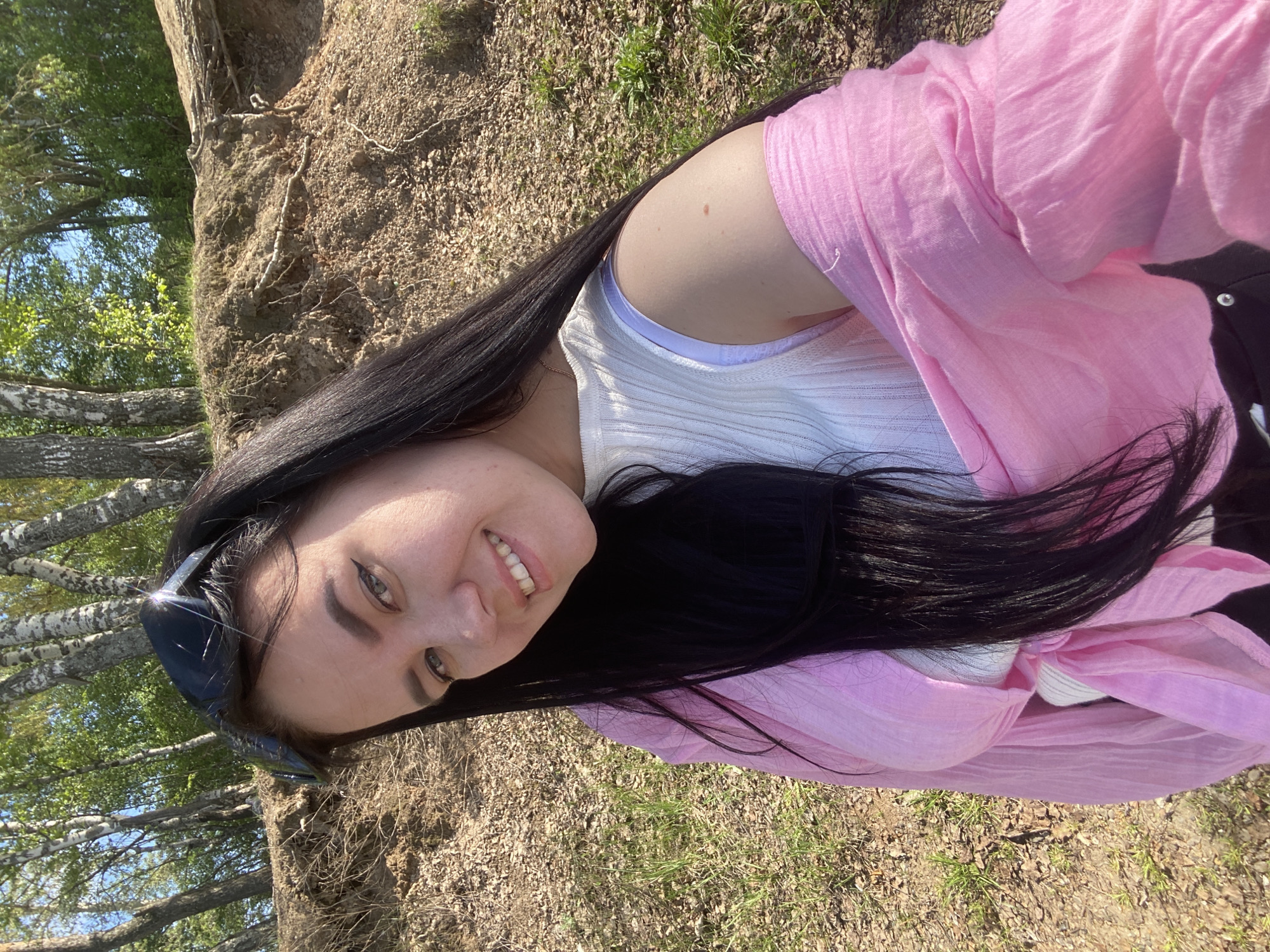 A woman is pictured in a selfie outdoors. She has dark hair, light skin, and is smiling. She is wearing sunglasses atop her head, a white tank top, and a pink shirt. The background includes trees, foliage, and a dirt slope. The location is Chaikovsky, Russia.