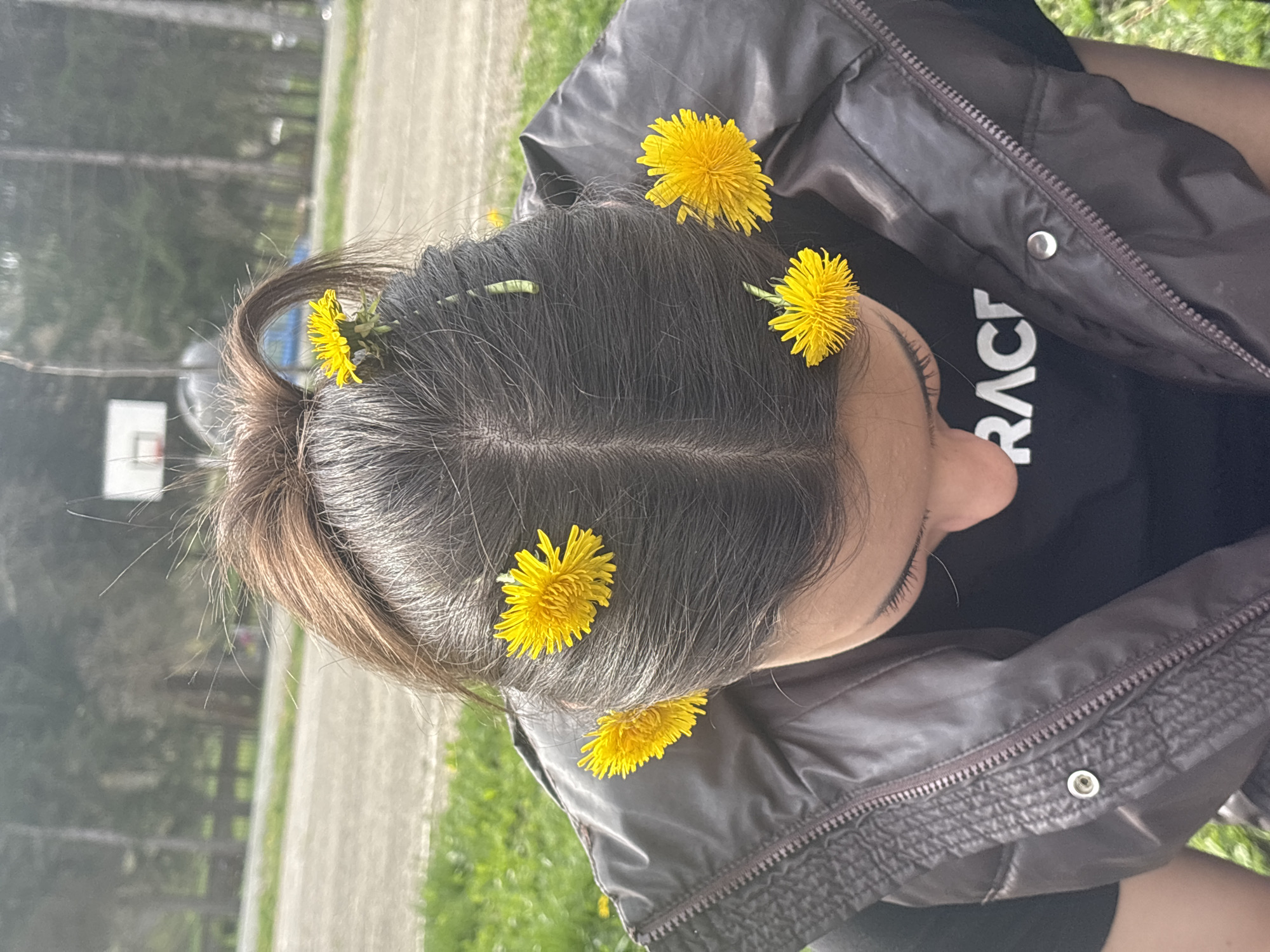 A woman is seen from an overhead perspective, seated on the grass. She has dark hair pulled back in a ponytail, adorned with several yellow dandelions. She is wearing a black t-shirt and a brown jacket. The background includes a grassy area, a paved path, and a basketball hoop. This photo was taken in the city of Troyan, Bulgaria.