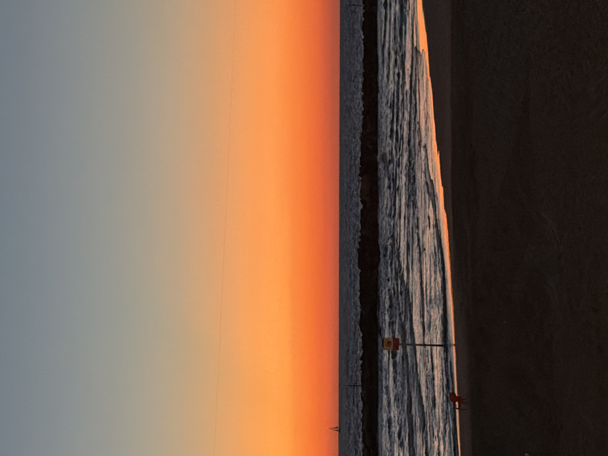 The image, oriented horizontally, captures a picturesque coastal scene at sunset. The sky occupies the upper portion, featuring a dramatic gradient from a pale, cool blue-gray on the left to a vibrant, warm orange on the right, indicative of the sun's low position.
Below the colorful sky lies a body of water, likely the sea, appearing dark blue-gray with subtle ripples and waves reflecting touches of the orange light. A dark, jagged line, consistent with a breakwater or a rocky shoreline, separates the sky and water.
On the far right, a dark strip of land or beach is visible, contrasting with the illuminated sky and water. A thin pole stands in the water near the breakwater, supporting a small, rectangular sign that appears to be red and white, though no text is legible. Very faint, dark silhouettes, possibly buoys or small boats, can be seen in the distance on the water.
The time of day is clearly sunset, creating a tranquil and visually striking atmosphere. No people are present in the scene. Given the context, this is a view of the Mediterranean Sea, likely near Tel-Aviv, Israel.