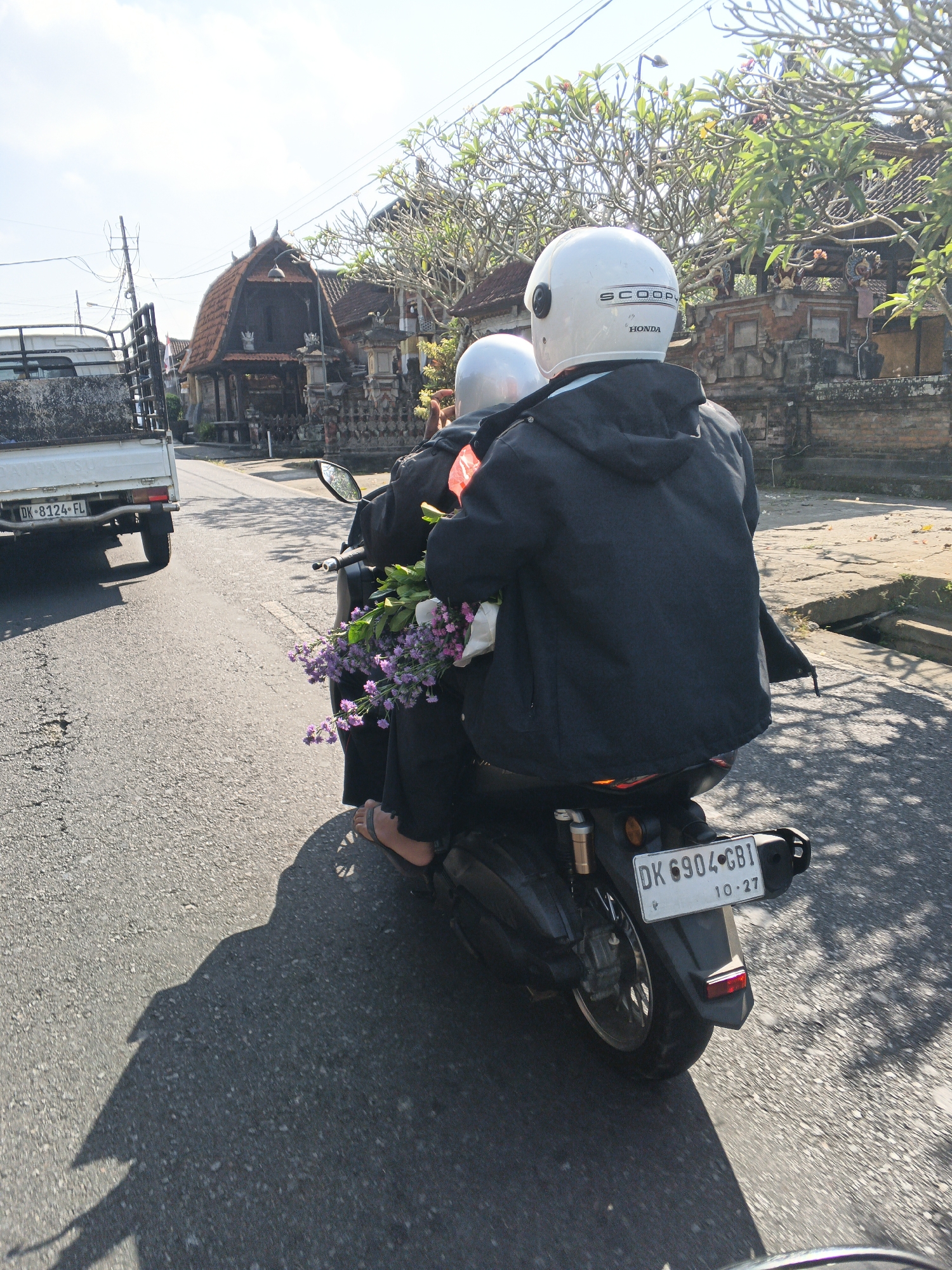 Two individuals are riding a scooter on a paved road in what appears to be the city of Abiansemal, Indonesia. The person in front is wearing a white helmet with "SCOOP" and "HONDA" branding, and a black jacket. The person behind them is wearing a lighter colored helmet and is holding a bunch of small purple flowers. A white truck is visible in the background on the left side of the road, with a license plate reading "DK 8124 FL". Traditional Balinese architecture, characterized by pitched roofs and ornate stone walls, lines the right side of the road. The ground is asphalt with visible cracks and patches, and shadows from trees and the scooter are cast across the road. The sky is bright and sunny. The scooter's license plate is visible and reads "DK 6904 GBI" with "10-27" below it.