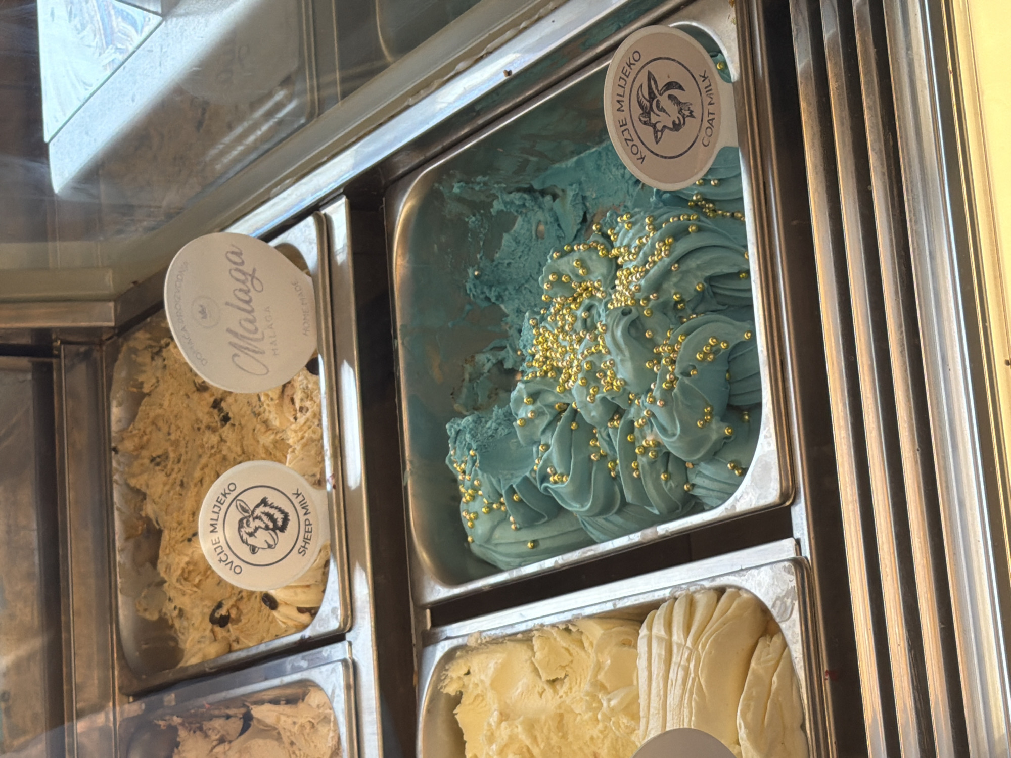 The image shows a close-up view of several metal containers filled with ice cream in a display case. In the foreground, a container holds light blue ice cream swirled and topped with small gold beads. To its left, another container holds pale yellow ice cream. To the left of that, partially visible, is a container with a light brown ice cream.
Two circular labels are visible. One, partially obscured, is on the container with the light brown ice cream, featuring a sheep's head in silhouette and text that includes "OVČIJE MLIJEKO" and "SHEEP MILK". The other label, on the container with the blue ice cream, also features a goat's head in silhouette and text that includes "KOZJE MLIJEKO" and "GOAT MILK". A third label, mostly visible in the upper right, is white and oval-shaped, with "Malaga" written in script, and "MALAGA" and "HOME MADE" in smaller print.
The display case appears to be part of an establishment selling ice cream. The lighting suggests an indoor environment, possibly a shop or gelateria. The background is slightly out of focus but shows more ice cream containers and the reflective surface of the display case. The overall impression is of a chilled display showcasing various flavors of ice cream. The location is not specified in the image itself, and no people are visible.