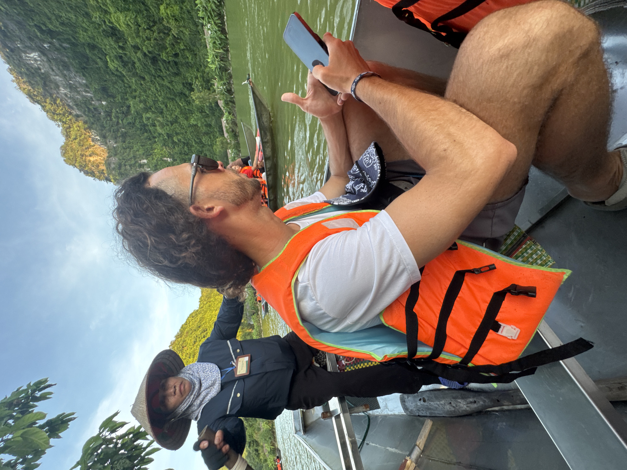 Two people are in a boat on a body of water, with a rocky, vegetated hillside in the background. The person in the foreground is a male wearing a white t-shirt, grey shorts, and an orange life vest. He has curly brown hair and is wearing sunglasses and a bracelet. He is holding a black smartphone in his right hand and gesturing with his left thumb. The person in the background is wearing a conical hat, a dark blue jacket, black pants, and a light-colored scarf around their neck. They are holding an oar, presumably steering the boat. The water is green, and other boats are visible in the distance. The sky is blue with some clouds, and the sun is illuminating the hillside. The environment appears to be a scenic natural setting, likely a river or lake in a mountainous region of Vietnam, possibly near Yên Sơn Ward, known for its karst landscapes and waterways.