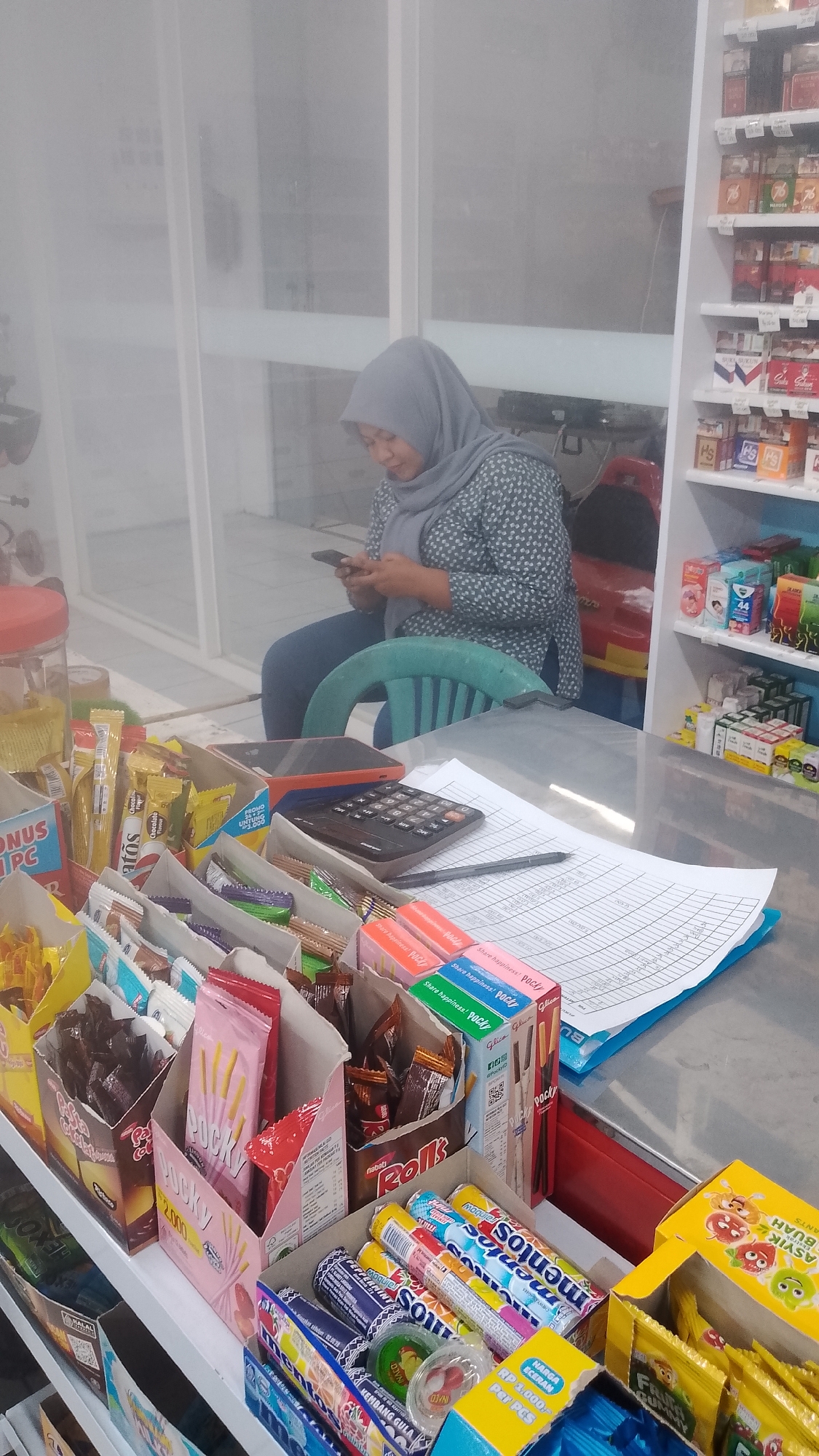 In a retail store located in Karanglangu, Indonesia, a woman wearing a grey hijab and a patterned blouse is seated and looking at her smartphone. In front of her, on a counter, is a black calculator and a pen lying on top of a document. The counter is lined with various snacks and candies displayed in cardboard boxes. To the right of the counter, shelves are stocked with more packaged goods, including cigarettes and toiletries. The store appears to be a small convenience store or minimarket, with bright lighting.