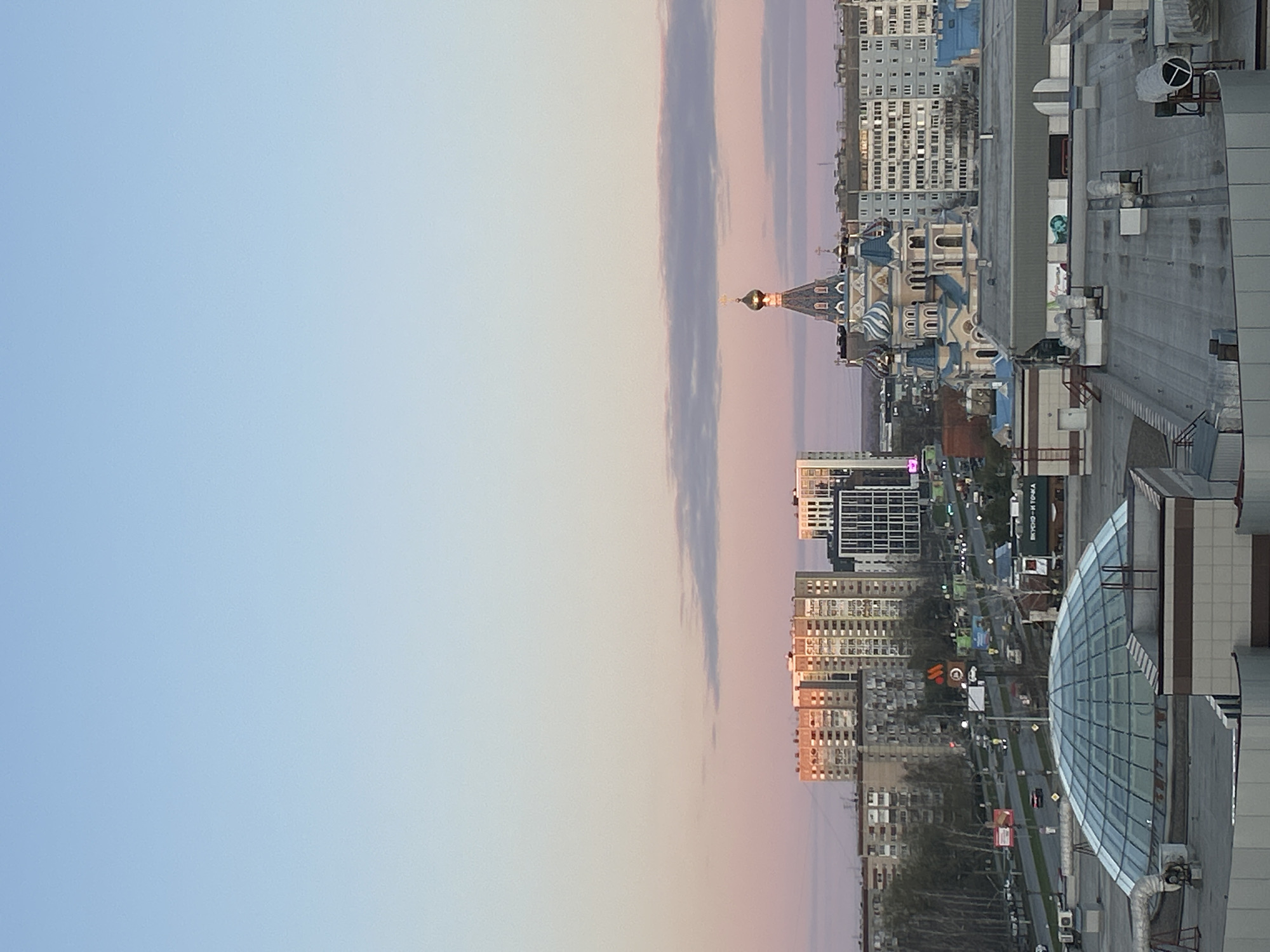 The image shows an urban view in Izhevsk, Russia, during what appears to be either sunrise or sunset. The sky transitions from a light blue to a pastel pink and orange. A strip of clouds runs vertically through the center of the frame. The cityscape includes a variety of buildings, ranging from residential blocks to modern structures with glass facades. A prominent, elaborately designed church with multiple domes is visible. A large building with a curved glass roof dominates the right side of the image. The scene appears to be photographed from an elevated position, looking out over the city. A highway runs parallel to the foreground buildings.