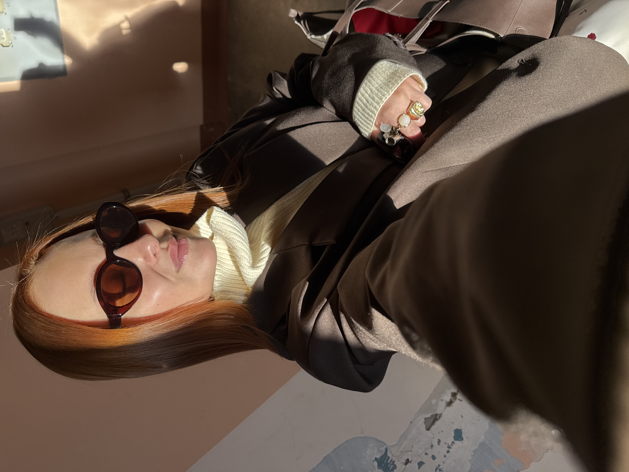 A woman with reddish-brown hair and brown sunglasses is wearing a brown blazer over a cream-colored turtleneck. She is wearing several rings on her right hand. Part of a brown bag is visible in the upper right. The scene appears to be indoors. There is no explicit evidence in the image to confirm the location in the city of Chaikovsky, Russia.