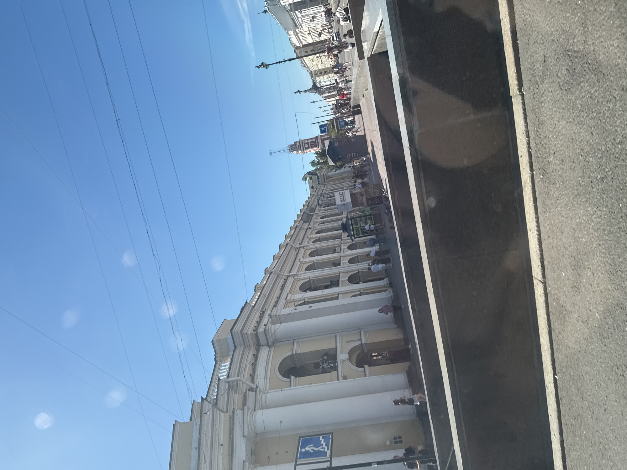 The image depicts a street scene in Saint Petersburg, Russia, likely near the Admiralty or Palace Square given the architectural style. In the foreground, a road is visible, with a raised concrete barrier and an asphalted surface. Beyond the barrier, a pedestrian walkway is lined with a building featuring a classical facade with columns and arched windows. A blue and white directional sign showing a person walking up stairs is mounted on a pole near the building. Several people are walking on the sidewalk. Further down the street, more buildings with white facades and columns line the right side, and a distinctive red and white tower with a spire rises in the distance. The sky is clear blue with some faint circular light reflections and overhead power lines. The overall impression is a bright, sunny day in an urban environment with significant historical architecture.