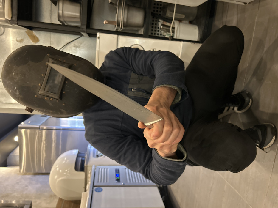 In a commercial kitchen located in Schiltigheim, France, an individual is observed in a deep squatting position. Their head is covered by a dark, worn welding mask featuring a small, rectangular viewing window secured by rivets. The individual is dressed in a dark blue long-sleeved fleece jacket with a lighter blue lining at the collar, dark athletic pants, and athletic shoes (one black with white stripes, the other black with grey accents). Both hands are clasped around the handle of a large, straight-bladed silver knife or cleaver, which is held vertically in front of their torso with the blade pointing upwards. The background on the left features white commercial kitchen appliances, including an ice machine with a blue digital display and a slotted vent, and a larger white refrigerator or freezer unit, along with stainless steel surfaces. On the right, a dark, multi-tiered shelving unit holds various metal containers and a white bucket. The floor is covered with light grey tiles.