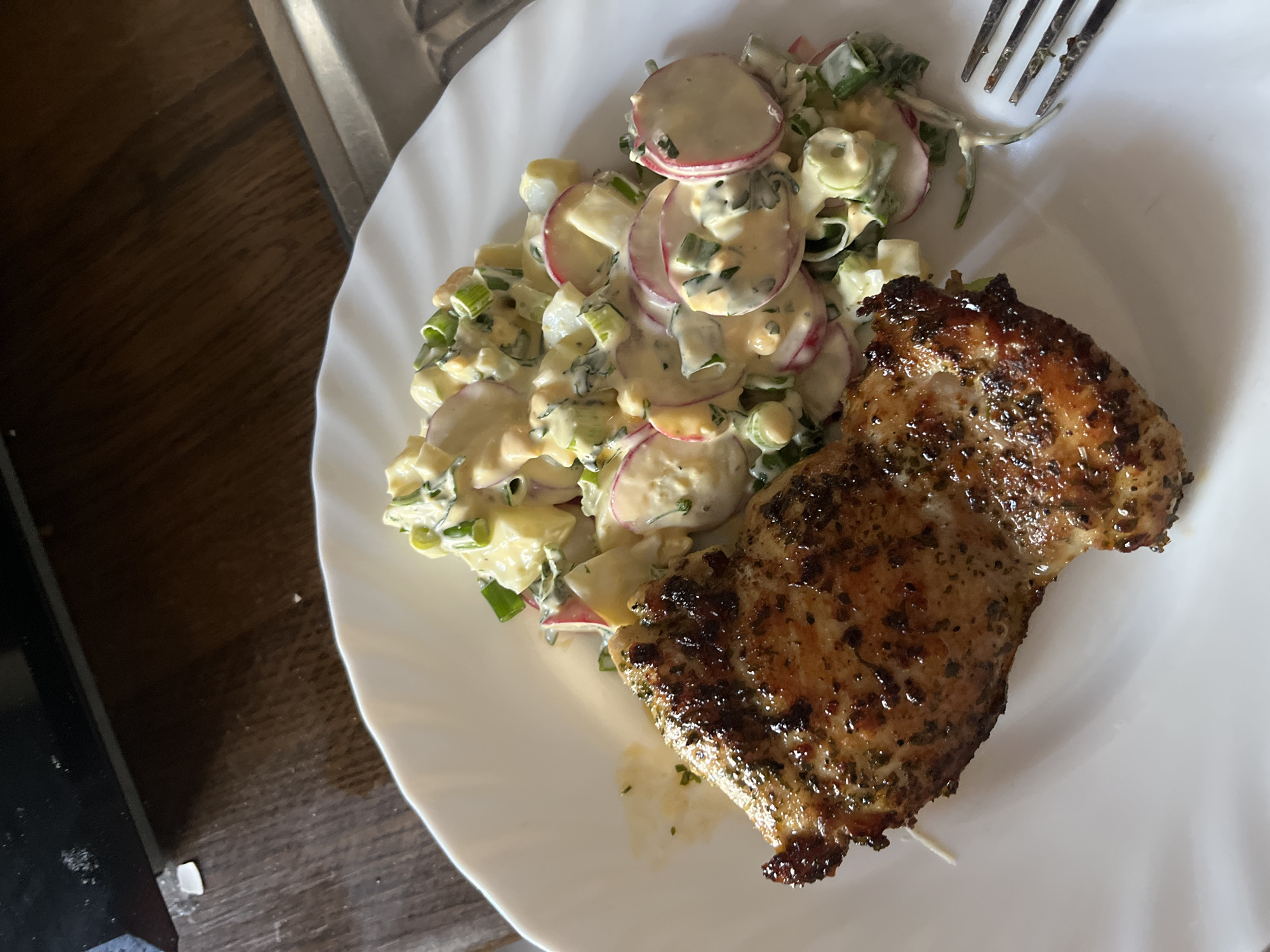 On a white plate, there is a piece of grilled meat next to a salad. The salad appears to contain radishes, potatoes, green onions, and a creamy dressing. A fork is also visible on the plate. The plate is sitting on a table, with a blurred surface visible in the background. The location is unknown.