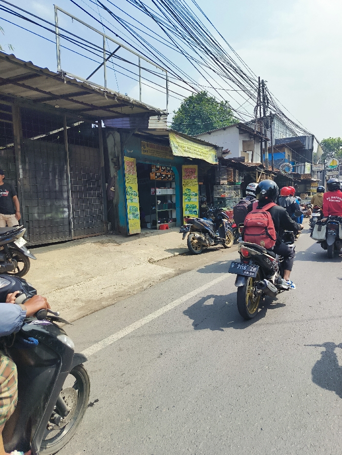 The image shows a street in Ngamprah, Indonesia (latitude: -6.858, longitude: 107.499). There are multiple motorcycles parked and driven along the road. Five people are present, four of them riding motorcycles and one standing on the sidewalk. The person standing is wearing a black shirt and a black helmet. The motorcycles are all black with red accents. The street is paved and has a white line down the center. The surrounding buildings are all single-story and made of brick. There are power lines strung across the street. The sky is blue and clear.