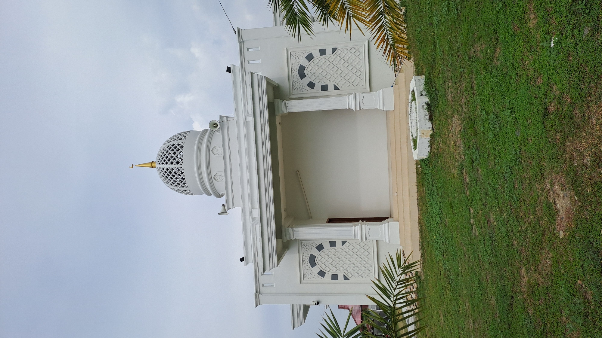 A white mosque with a golden dome and a crescent moon sits in the city of Bireuenmeunasahdayah, Indonesia. The mosque has a modern design with clean lines, a large covered entryway supported by columns, and decorative geometric patterns on its facade. Speakers are mounted on the sides of the building, and palm fronds frame the lower portion of the image. A grassy lawn surrounds the mosque, with a small concrete planter in the foreground. The sky is overcast, with a few clouds visible.