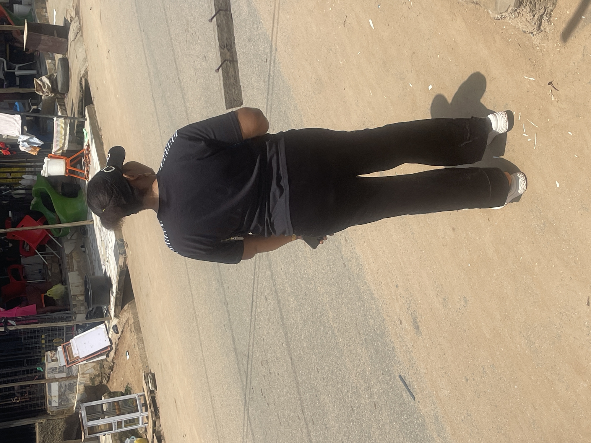 A person wearing a black t-shirt, black pants, a black baseball cap, and white sneakers is standing on a dusty, paved road, facing away from the camera. They are holding a dark object, possibly a phone, in their right hand. The person's shadow is cast on the ground to their right. In the background, there is a row of market stalls or shops. One stall features a display with a glass front, showing some items inside. Other stalls have various goods, including what appear to be red chairs, plastic containers, and some textiles. A green plastic slide is visible in the background. The overall environment suggests a commercial area in a city, likely Abuja, Nigeria, with a mix of commercial activity and open ground.