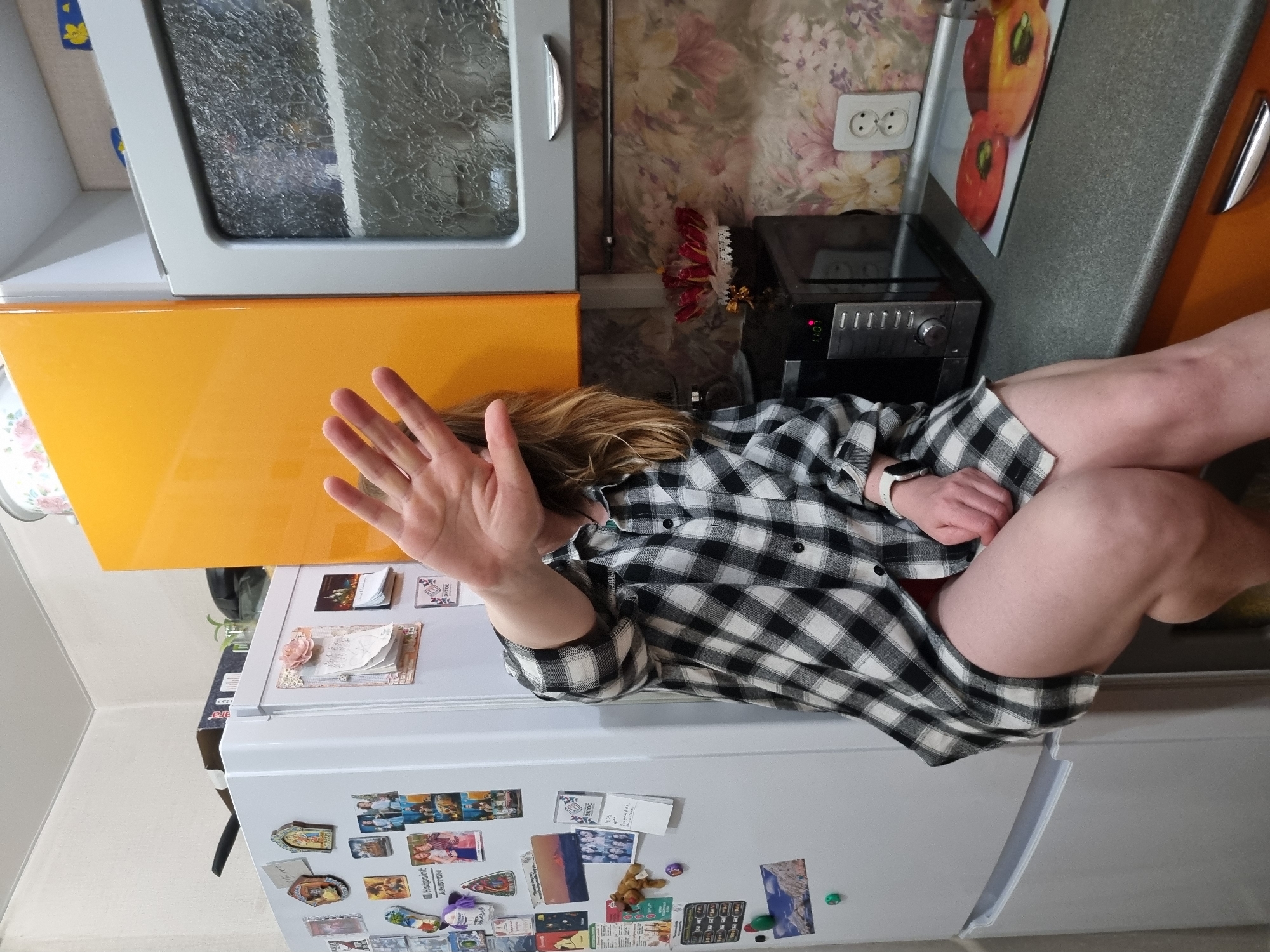 A person sits on a white refrigerator in a kitchen. The person is wearing a black and white checkered shirt and a smartwatch. Their legs are bare. The person raises their left hand as if to obscure the camera.
The refrigerator has magnets and photos affixed to it. Above the refrigerator is a yellow cabinet and a gray cabinet. A small black microwave oven is to the right of the person, next to a wall with floral wallpaper and a European-style electrical outlet.
While the image includes details suggestive of a domestic kitchen environment, there is no explicit information within the image to confirm its location in the city of Chaikovsky, Russia, or any other specific location.