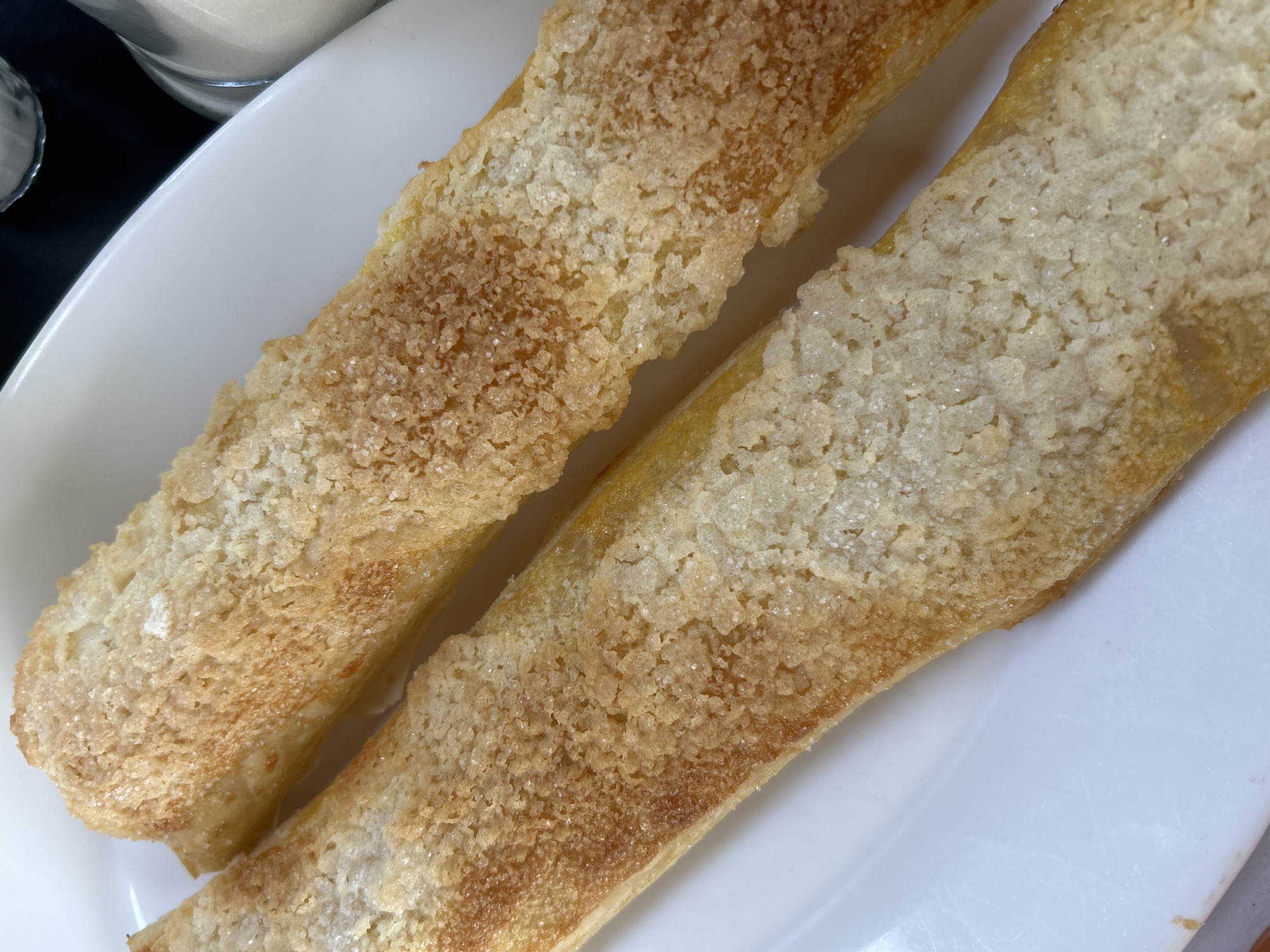 Two pastries covered in sugar sit on a white plate. The pastries are long and rectangular, appearing golden brown with a textured, sugary surface. The background is blurred, revealing a glass container and dark surface. The photo was likely taken indoors. The location and city are not determinable from the image.