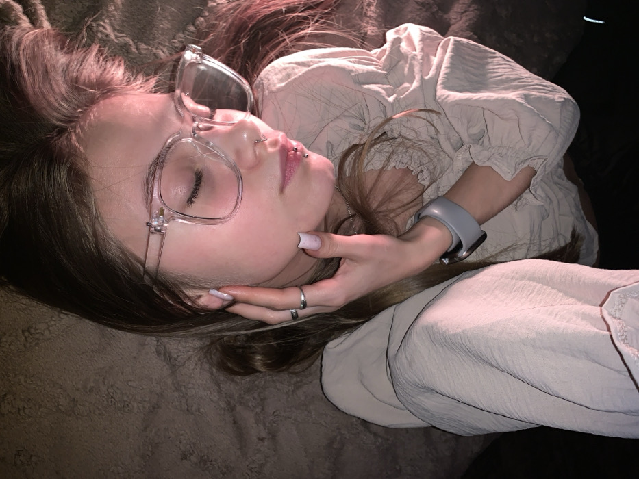 A person is depicted lying on a textured, brownish-gray surface, likely bedding, in an indoor setting in Фоки, Russia. The subject's head is tilted right, with eyes closed, illuminated by a warm pinkish light. They wear clear-framed glasses and have multiple facial piercings, including a double piercing on the upper lip and two on the lower lip. The person's left hand, adorned with two silver-colored rings and light-colored manicured nails, rests on the left side of their face. A gray smart watch is visible on the left wrist. The subject is wearing a light beige, ruffled blouse or top, with another similarly colored garment visible on the right. Long, light brown hair is spread around the head, and a thin silver chain is visible around the neck. The background is dark, suggesting a low-light environment.