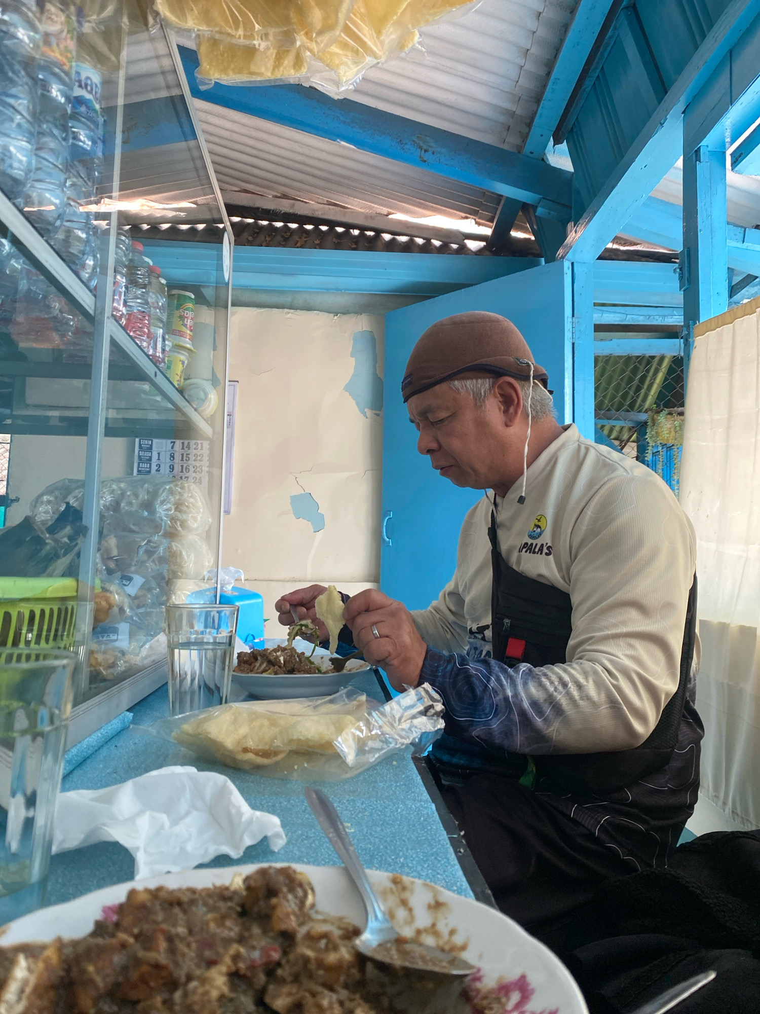 A middle-aged Asian man with a brown cap is eating a meal at a blue table. He is wearing a light-colored long-sleeved shirt with a black fanny pack. In front of him are two plates of food, a glass of water, and a stack of crackers in a plastic bag. The background shows a shelf with bottles of water and snacks. The scene appears to be an outdoor food stall or a casual eatery.