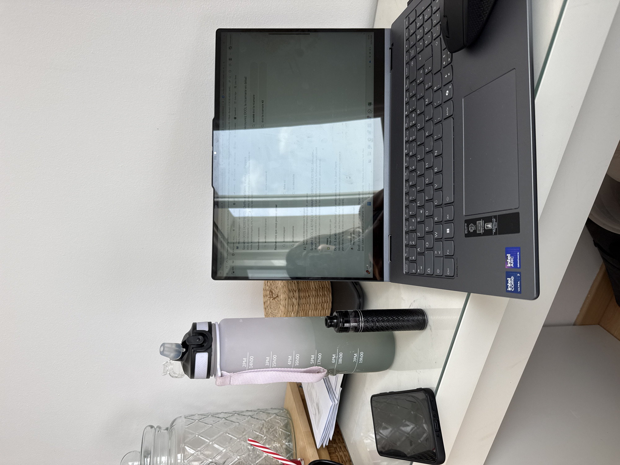 A laptop, water bottle, vape, and phone are on a white desk in an indoor setting. The laptop is open and displays a search for "DCCL la ricamarie." "La Ricamarie" is a city in the Loire department in central France. The water bottle is grey and marked with hours. A black vape pen is next to the water bottle. A black smartphone is lying on the desk. The background includes a white wall and wooden shelving.