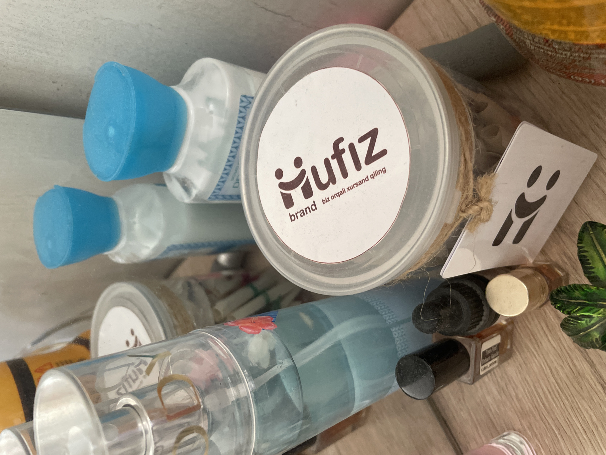 The image depicts a collection of cosmetic and fragrance products arranged on a wooden surface. In the foreground, there are several small bottles, including what appears to be nail polish and an oil dropper. A larger, clear bottle with a blue liquid and a spray top is prominently displayed on the left, with a floral design on its side. To the right of this bottle is a transparent container with a white lid that has a logo and text that reads "iufiz brand biz orqali xursand qiling." Behind these items, two white bottles with bright blue caps are visible, suggesting they are part of a personal care line. The background shows a textured wall and reflections in a mirrored surface, indicating the products might be displayed on a vanity or dresser. The overall scene is consistent with a retail or personal collection of beauty products. The reference to Tashkent, Uzbekistan, is not directly evident from the visual information within the image.