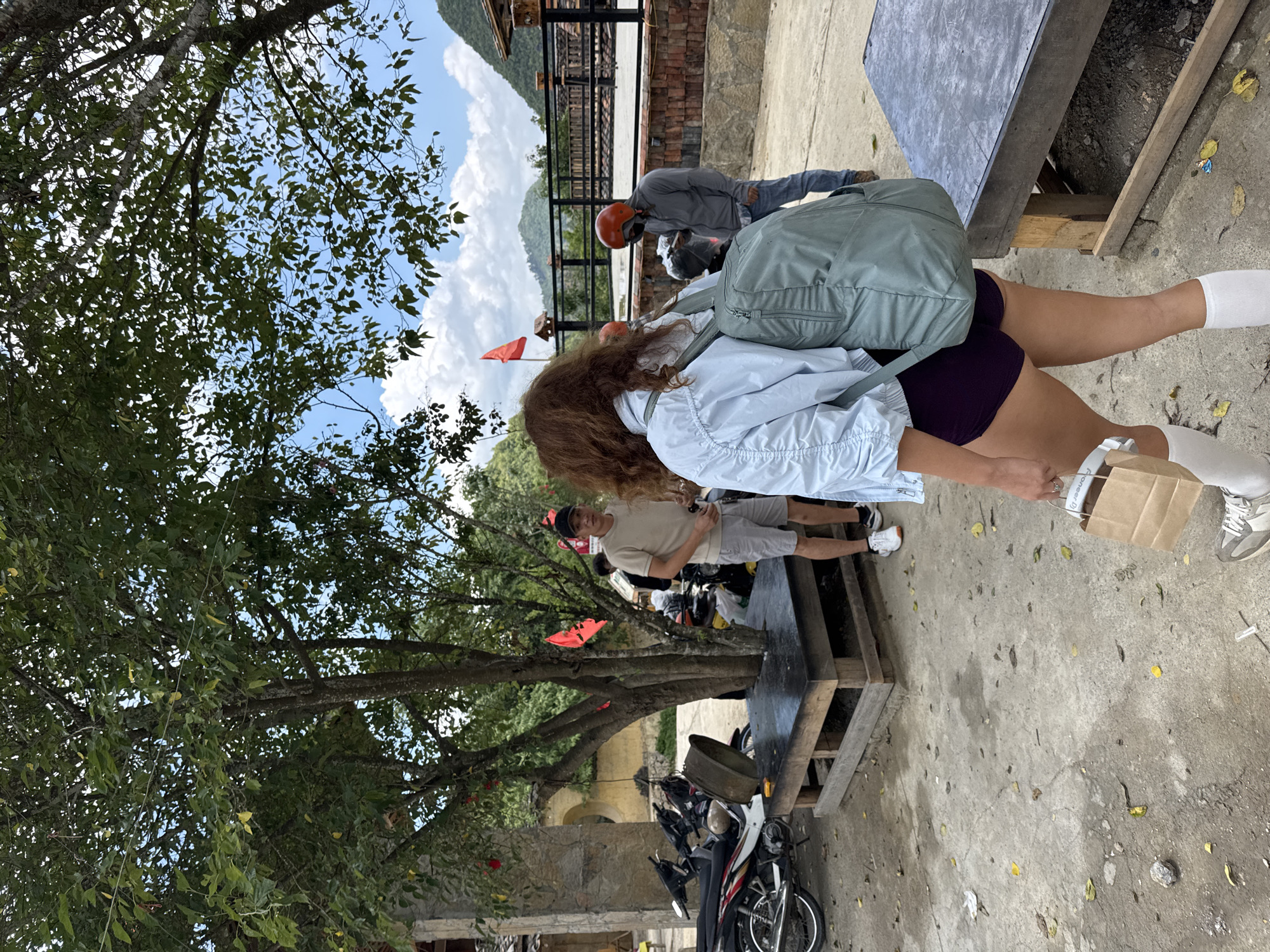 The image, taken in Lũng Cú Commune, Vietnam, captures an outdoor scene. A person with long, reddish-brown hair, dressed in a light blue shirt, dark shorts, and white socks, stands on a concrete surface with a light green backpack. They also hold a brown paper bag in their right hand. Behind this individual, another person in a gray shirt and shorts stands near a motorcycle parking area. An orange helmet is visible on one of the motorcyclists. A third individual wearing a gray jacket and orange helmet is seen near a brick structure. A rustic wooden table with a dark gray top stands in the foreground. A large tree with lush green foliage dominates the left side of the frame. In the background, mountains and a cloudy sky are visible. Red flags are placed in the area. Motorcycles are parked haphazardly. The scene conveys a sense of casual activity within a rural or semi-urban environment.