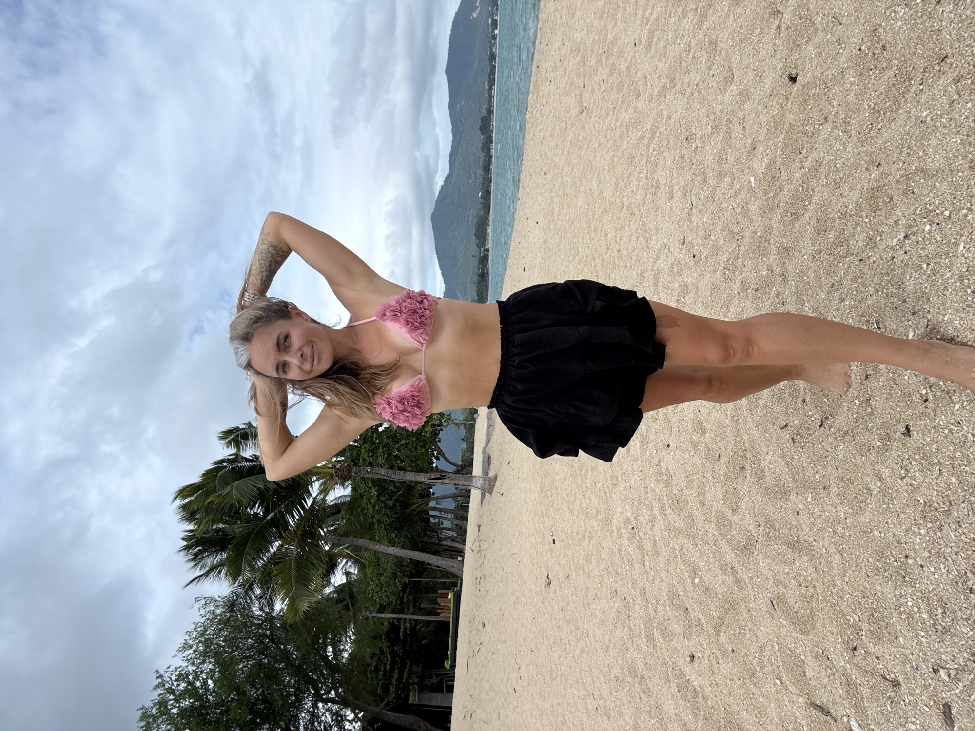 A woman with blonde hair and gray streaks stands on a sandy beach in the Ko Samui District, Thailand. She is wearing a pink floral bikini top and a black ruffled skirt. Her hands are raised behind her head, and she is looking at the camera with a slight smile. In the background, palm trees, lush vegetation, and mountains are visible against a cloudy sky. The ocean stretches out to the horizon.