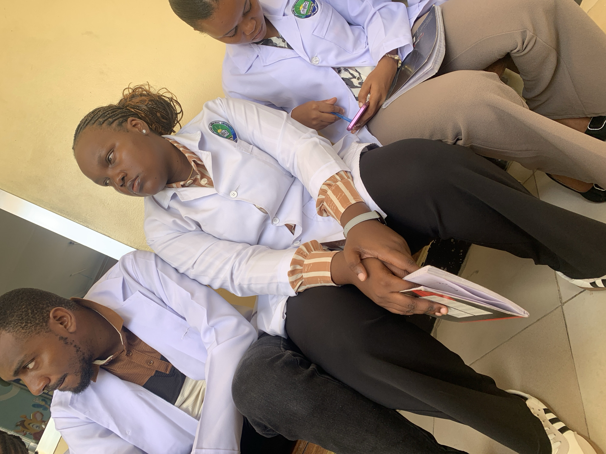 Three individuals, two women and one man, are depicted in a close-up, angled shot, seated against a light yellow/beige wall on a tiled floor. All three are wearing white lab coats, suggesting a medical, scientific, or educational setting.
The central figure is a woman with braided hair pulled back. She wears her white lab coat over a striped brown and white long-sleeved shirt with ruffled cuffs, black trousers, and white sneakers with black stripes. She holds a small, open notebook or booklet in her hands and looks to her right with a pensive or slightly tired expression. A patch on her lab coat clearly displays a crest and the text "MEMORIAL UNIVERSITY."
Behind her, towards the upper right, another woman is focused on a pink smartphone, using a blue stylus. She is also dressed in a white lab coat worn over a patterned top, light brown trousers, and black flat shoes. A watch adorns her wrist. Her lab coat also features a "MEMORIAL UNIVERSITY" patch.
To the lower left, a man with a short beard is seated, looking down and to his left, appearing contemplative. He wears a white lab coat over a brown collared shirt with a dark central panel, and dark trousers.
The scene appears to be set indoors or in a semi-enclosed area, with bright ambient light suggesting daytime. The individuals seem to be resting or waiting, possibly during a break or between activities at a university or college in Dar es-Salaam, Tanzania, given the visible text and location context. Their expressions range from focused to thoughtful, contributing to a quiet, reflective atmosphere.
