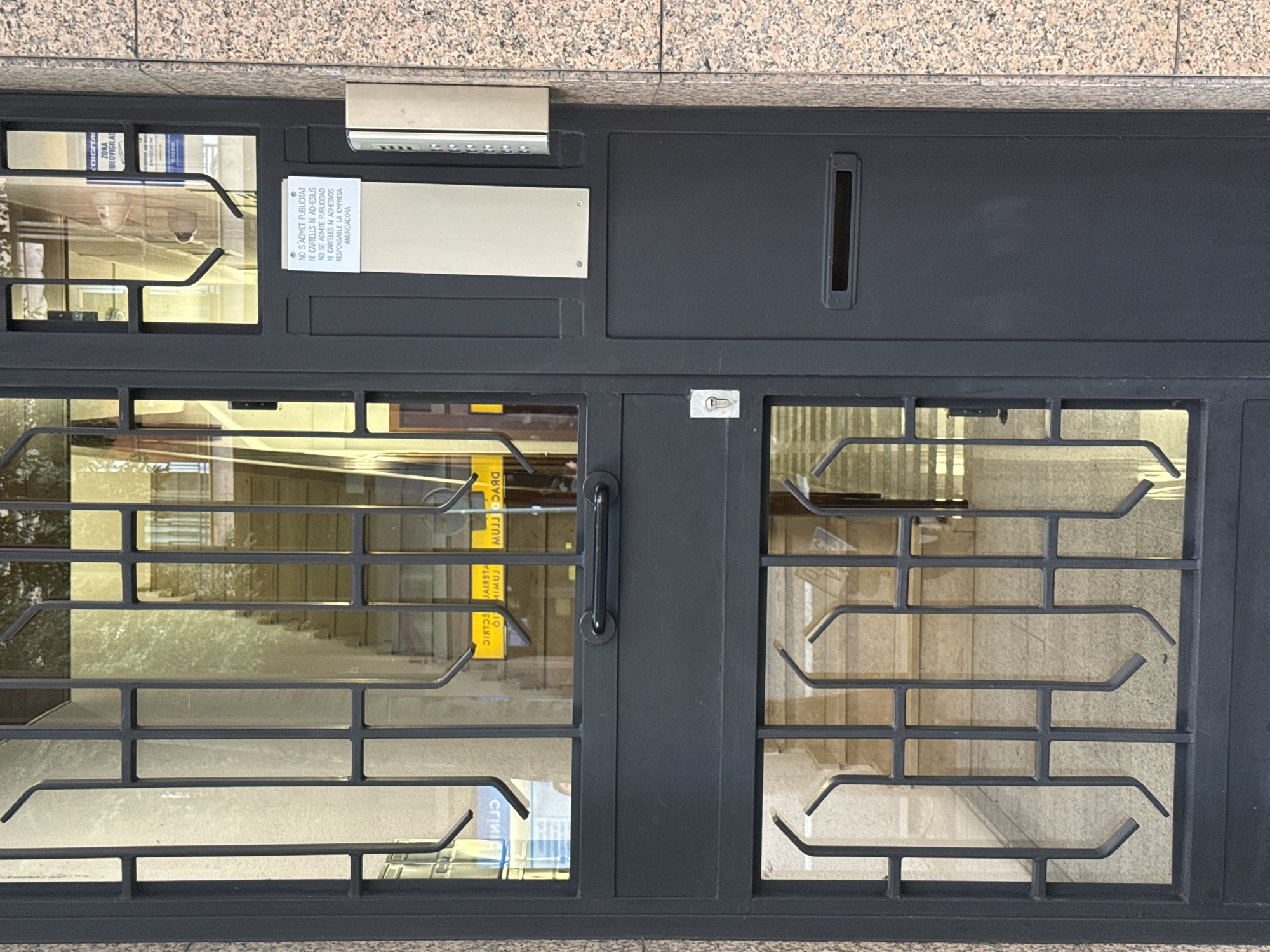 This is a medium-shot, eye-level, outdoor, eye-level, full-shot photograph of a dark gray metal door with a metal gate over a window. The door is on the first floor of a building, and it is set into a wall covered with speckled pink and gray granite. The door has a mail slot and a door handle. The gate is composed of thin metal bars. The window is made of glass, and it looks out onto a staircase. There is a small white sign attached to the door, with text printed on it in black. The text reads: "NO S'ADMÉT PUBLICITAT NI CARTELLS NI ADHESIUS. NO SE ADMITE PUBLICIDAD NI CARTELES NI ADHESIOS RESPONSABLE LA EMPRESA ANUNCIADORA." A silver colored intercom system is mounted to the wall next to the door. This is likely the entrance to a building in Barcelona, Spain.