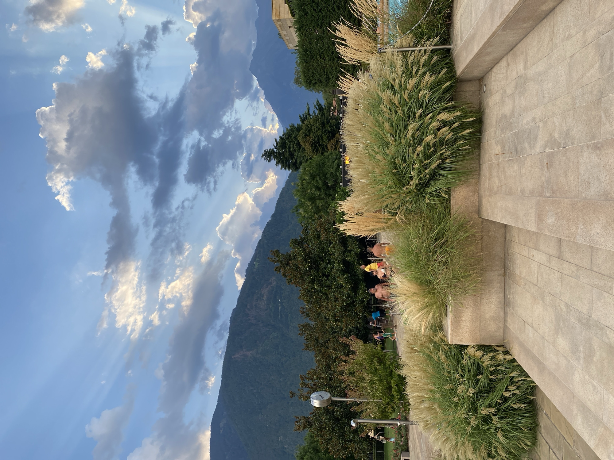 The image depicts a scene at the Meran - Merano thermal baths in Italy, likely during the late afternoon or early evening, judging by the lighting. The foreground is dominated by a paved area with light-colored stone tiles, leading up to a raised planter bed filled with tall, feathery ornamental grasses. In the middle ground, several people are visible, some sitting and others standing, appearing relaxed. They are casually dressed, with some wearing swimwear, suggesting they are enjoying the facilities. Behind them, lush green trees form a dense backdrop, with a clock mounted on a pole visible to the left. Further in the background, majestic mountains rise, their slopes covered in dark green vegetation, with dramatic clouds casting shadows and allowing shafts of sunlight to break through, creating a striking visual effect in the sky. The overall atmosphere is tranquil and scenic, showcasing the natural beauty surrounding the thermal baths.