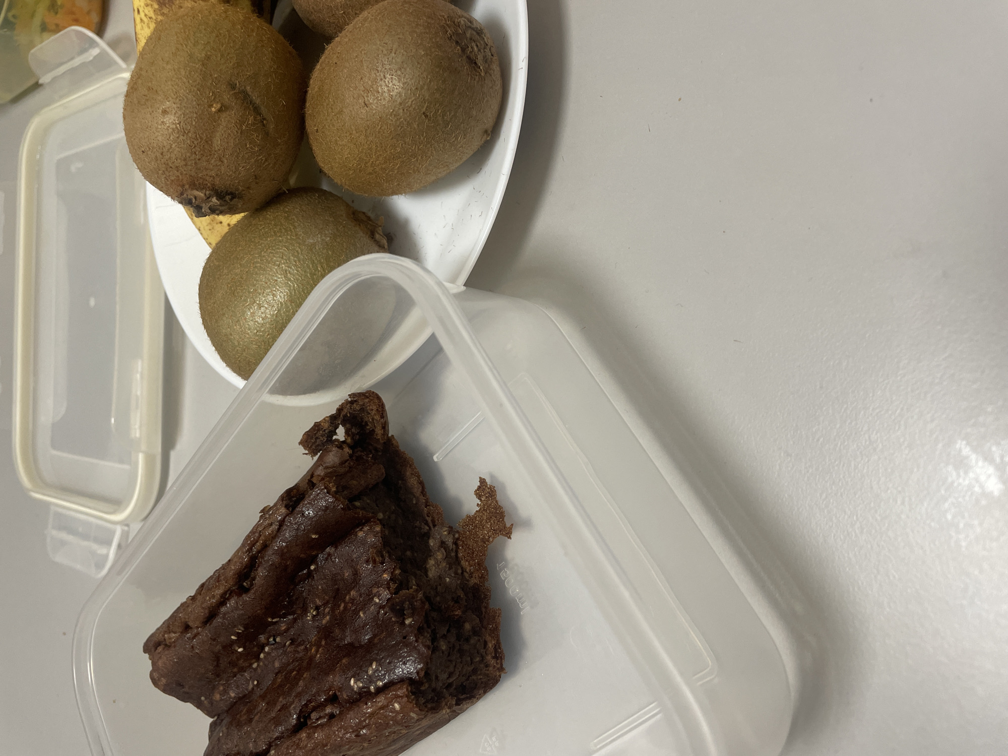 A slightly overhead shot captures a still life on a light grey surface. The scene contains food items: four kiwis are grouped together on a white plate, partly obscuring a banana underneath. A portion of what appears to be a brownie is placed inside a clear, rectangular plastic container. The lid of another identical container lies beside the plate of fruit. The location is not discernible from the image, making it impossible to confirm if it is Sofia, Bulgaria.
