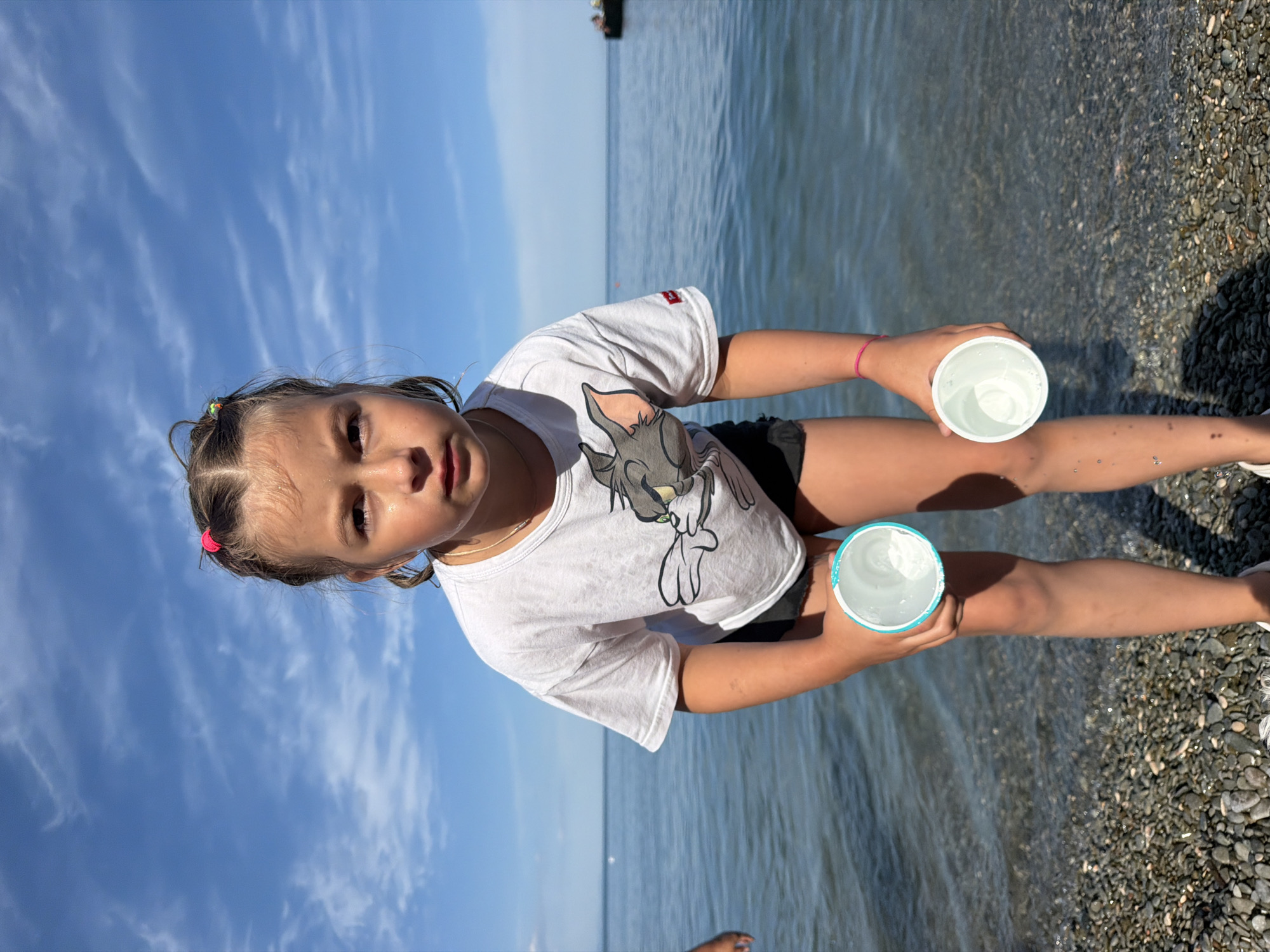 A young girl with brown hair, tied back with a pink hair tie, sits by the water. She is wearing a white t-shirt with a cartoon character printed on it and black shorts. In her hands, she holds two white cups, one in each hand. The girl is looking towards the camera with a curious expression.
The scene is set at a beach with calm water and a pebbly shore. The sky is blue with wispy white clouds, suggesting a clear, sunny day. The lighting indicates it is likely daytime, possibly late morning or afternoon. In the distance, a dark silhouette of a boat is visible on the water. The overall atmosphere is relaxed and summery, with the girl appearing to be enjoying her time by the sea.
There is no visible text in the image. The provided location context is Partenit, Ukraine.